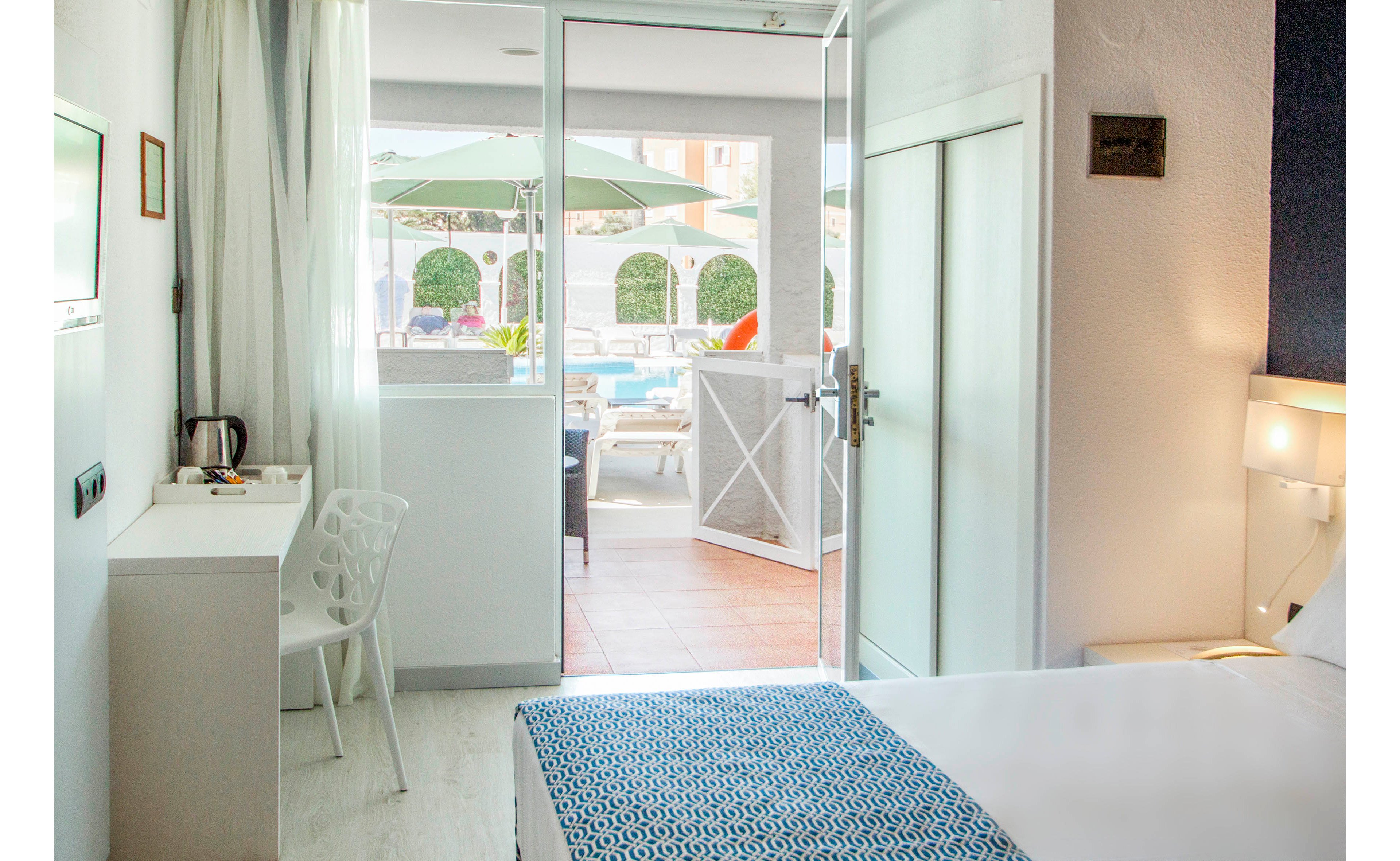 A modern hotel room features a neatly made bed with a patterned blue blanket and a small white desk with a chair and electric kettle. Sunlight streams through the open glass door leading to an outdoor terrace. Outside, green umbrellas and lounge seating surround a swimming pool, suggesting a relaxing vacation setting. The overall atmosphere is clean, airy, and inviting with light-colored furnishings and walls.
