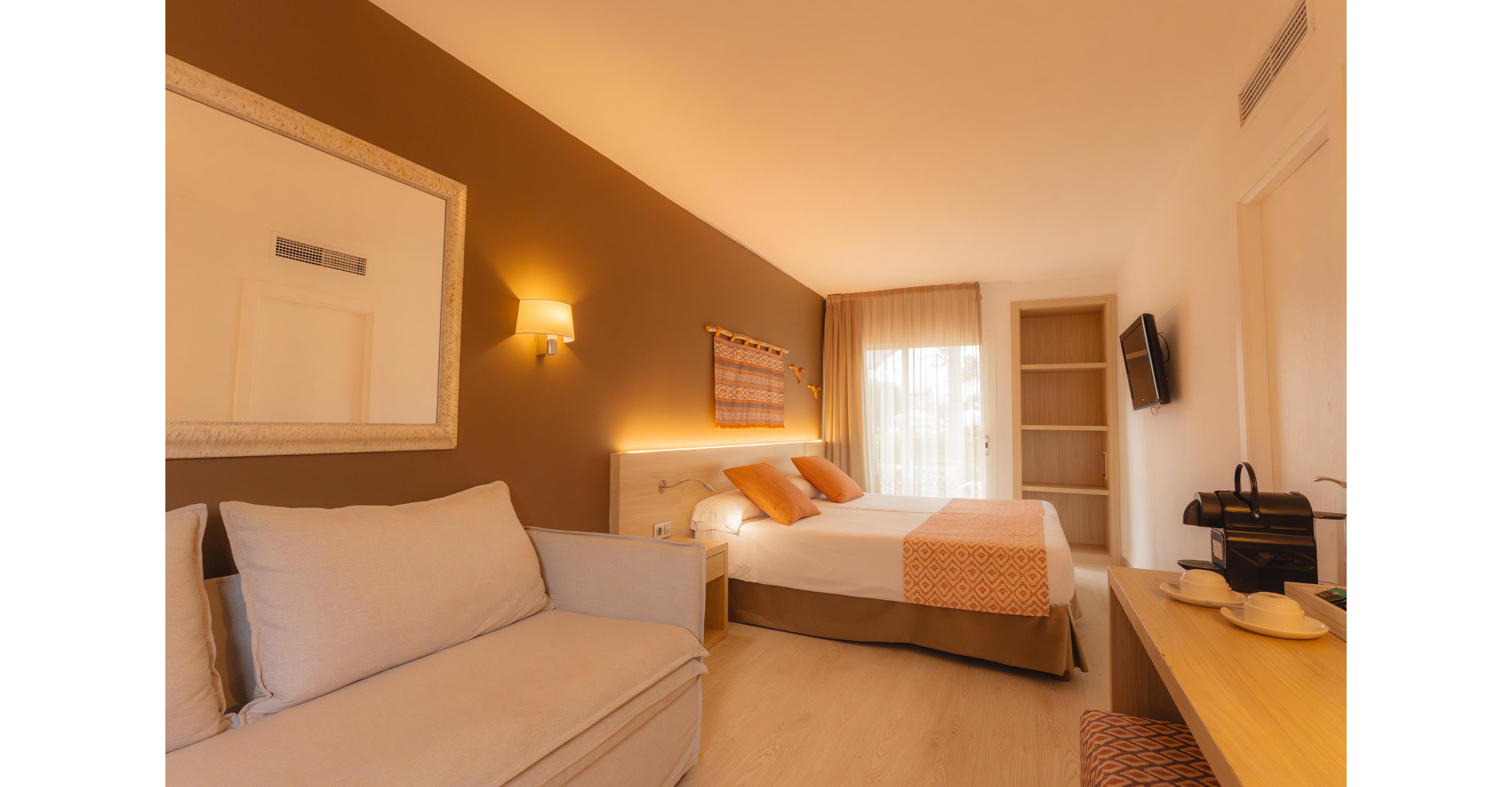 A contemporary hotel room features a neatly made double bed with orange accent pillows and runner against a brown feature wall. A light-colored sofa sits in the foreground, facing a wall-mounted television and a wooden desk with a coffee machine and cups. Soft warm lighting from a wall sconce and natural light through sheer curtains create a welcoming atmosphere. The overall design is minimal, with neutral tones and subtle orange details adding a touch of color.