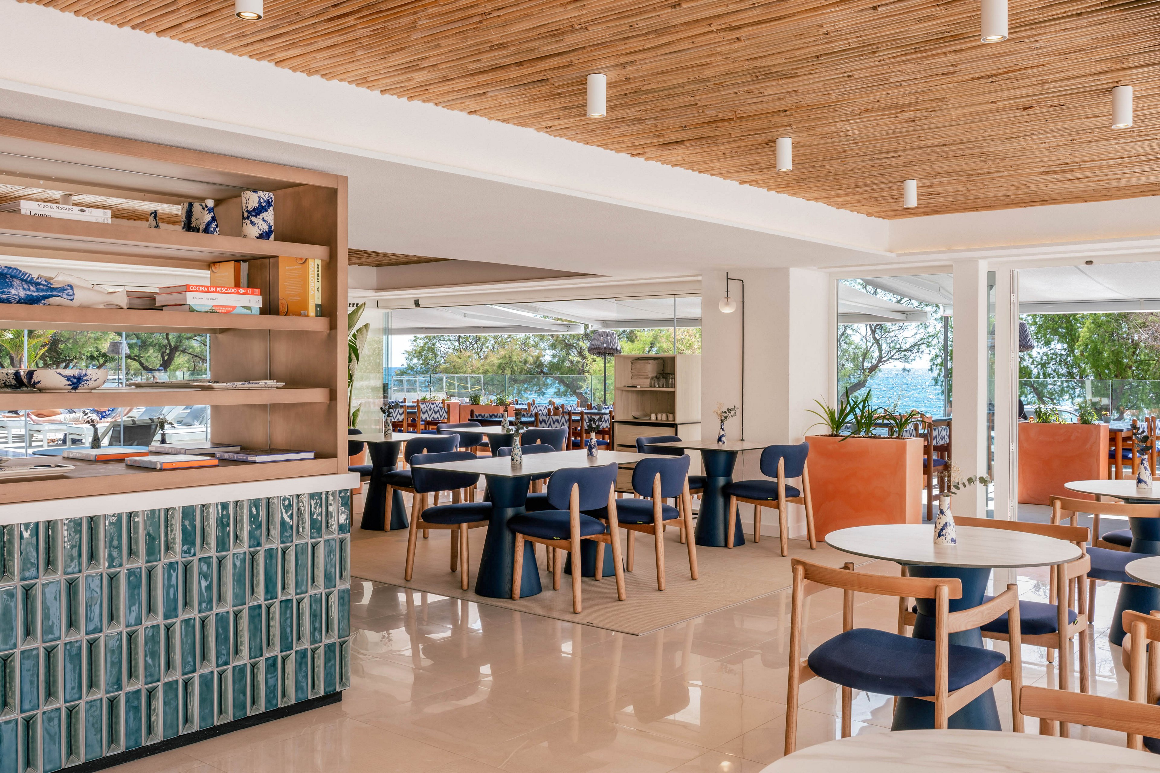 A modern restaurant interior features wooden chairs and tables with blue seat cushions and tan leather backs. Large windows and open walls reveal greenery and a glimpse of water outside, suggesting a coastal or lakeside setting. The space includes a tiled counter with vertical blue-green tiles and open shelving with decorative objects. Warm natural light, a wooden slat ceiling, and neutral flooring create an airy, relaxed dining atmosphere.