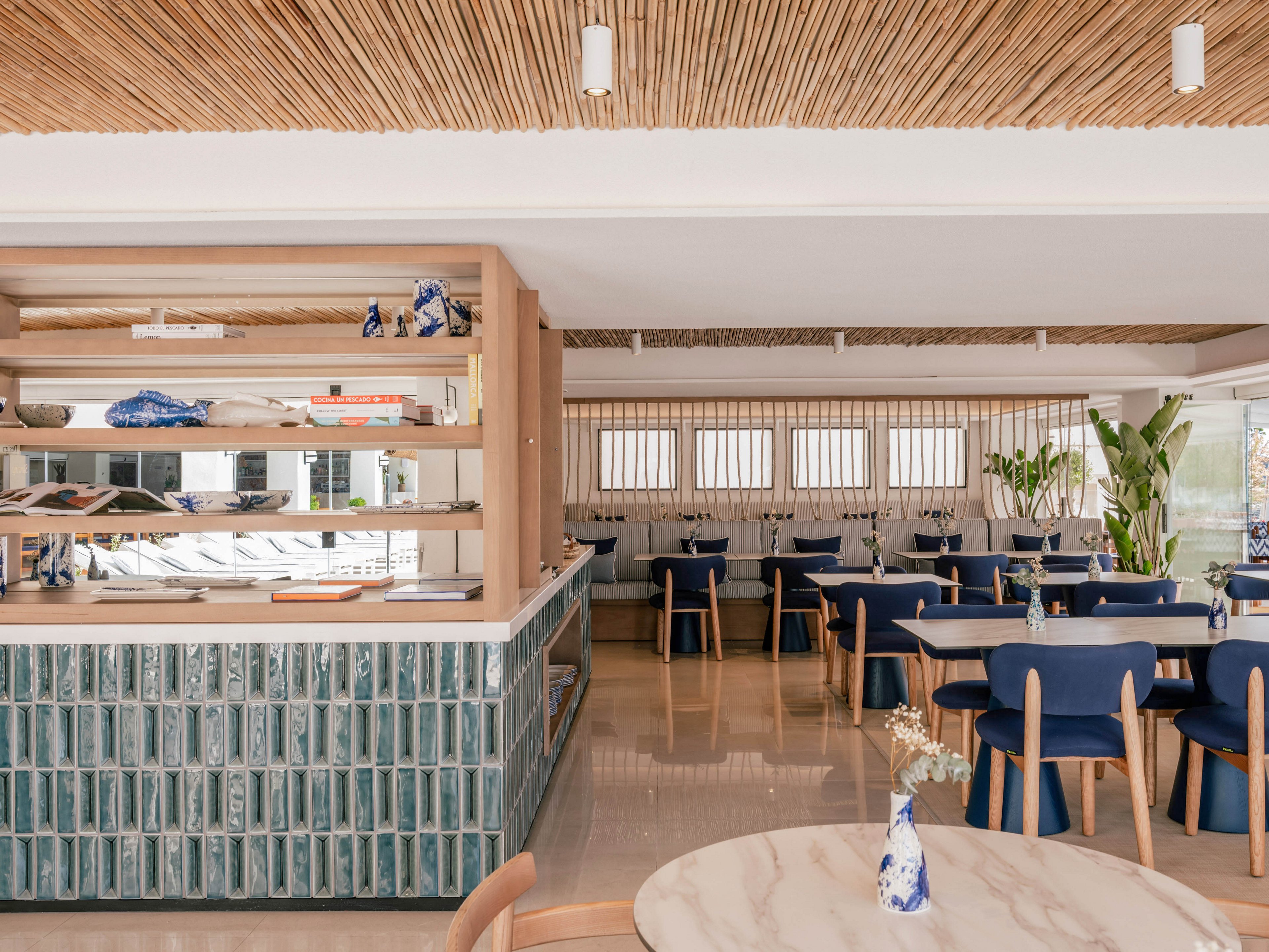 Contemporary restaurant interior featuring blue upholstered chairs and light wooden tables. A tiled counter with vertical greenish-blue tiles and open wooden shelving divides the space. Large windows and light-colored flooring create an airy, sunlit atmosphere. Decorative plants, ceramics, and dried flowers add a relaxed, coastal-inspired touch.