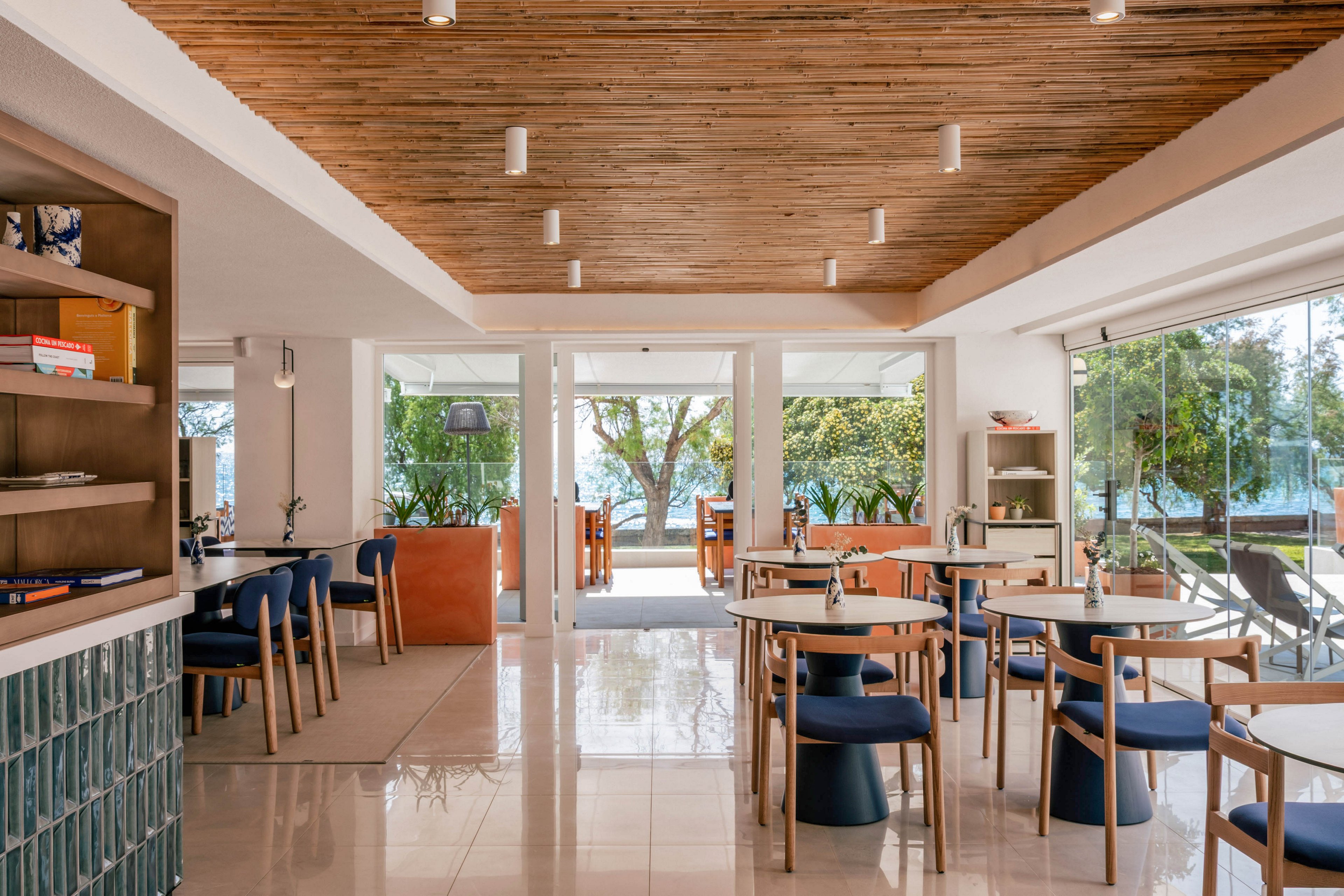 A contemporary cafe interior features wooden tables and blue cushioned chairs arranged neatly on a glossy tiled floor. Large glass doors open to an outdoor seating area with trees and greenery visible in the background. The ceiling is finished with warm wooden slats and minimalist white light fixtures. Natural light fills the space, creating an inviting and relaxed atmosphere suitable for dining or casual meetings.