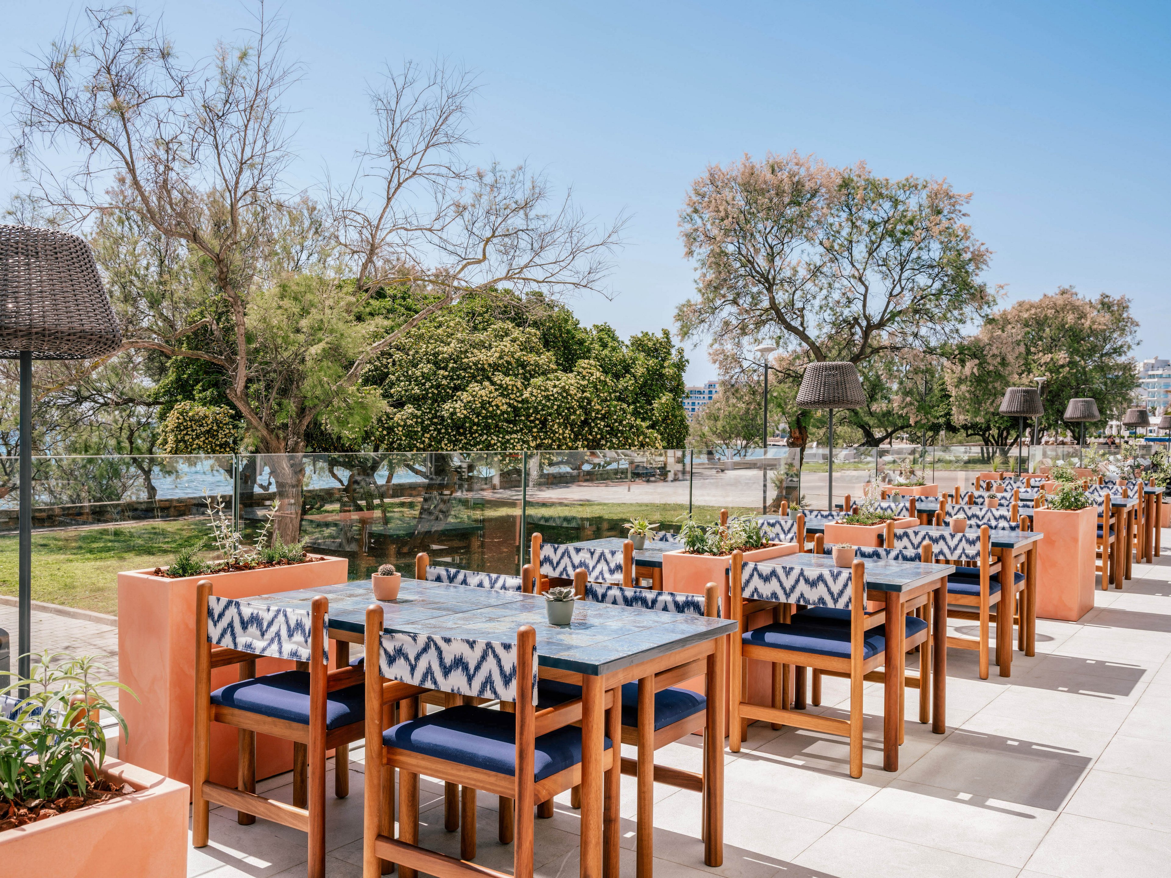 An open-air restaurant terrace features rows of wooden tables and chairs with blue cushions and chevron-patterned backs. The seating area is bordered by low planters and glass railings overlooking a green park and distant water. Tall outdoor heaters or lamps stand between the tables, adding structure to the scene. Bright daylight and clear blue skies create a relaxed, summery atmosphere.
