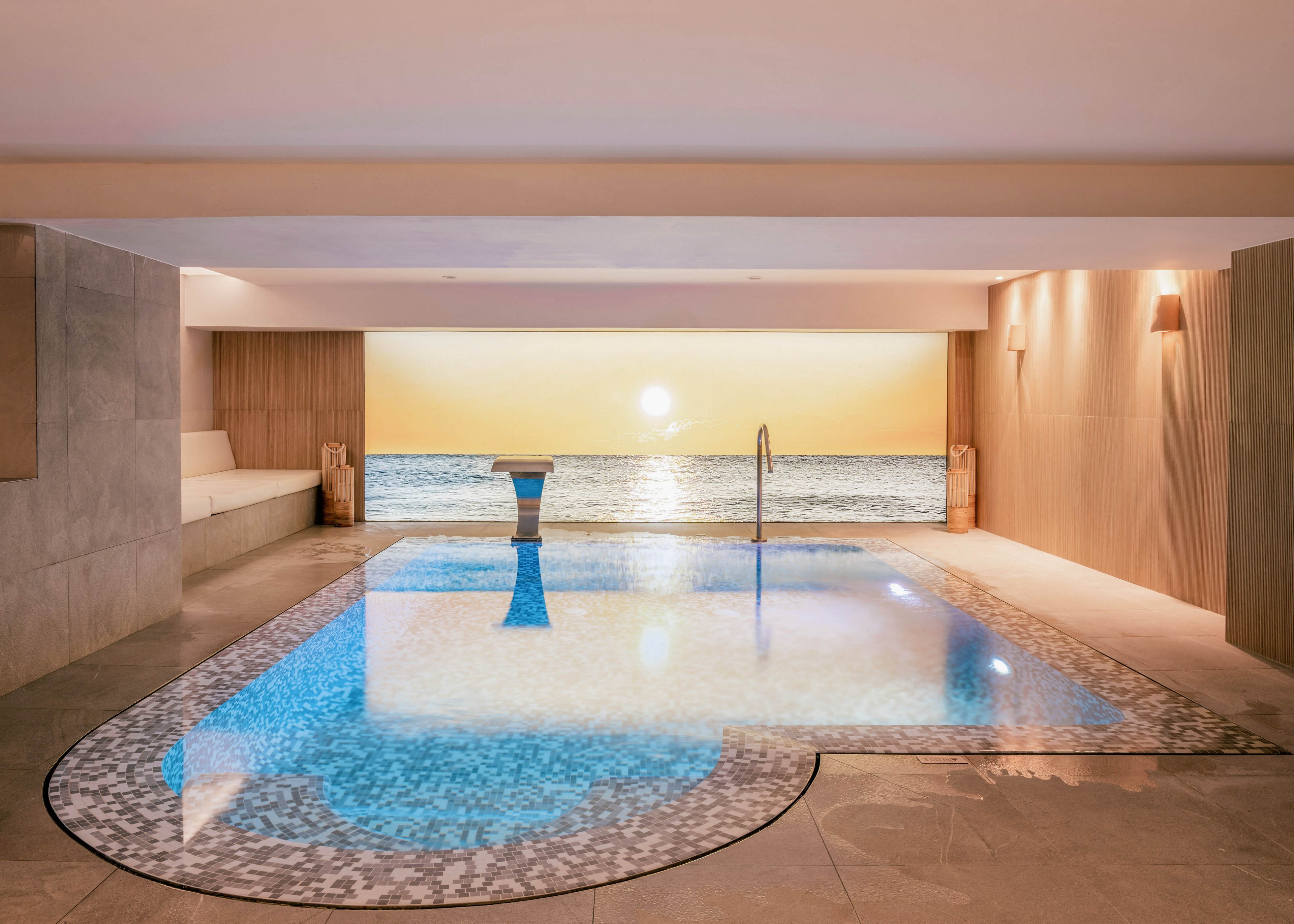 A modern indoor swimming pool is featured in a minimalist spa-like room. The pool has a mosaic-tile border and a central metal handrail leading into the water. At the far end, a large window or digital wall displays a serene ocean horizon with the sun setting over calm waves. Soft, warm lighting and built-in seating areas create a tranquil, upscale atmosphere.