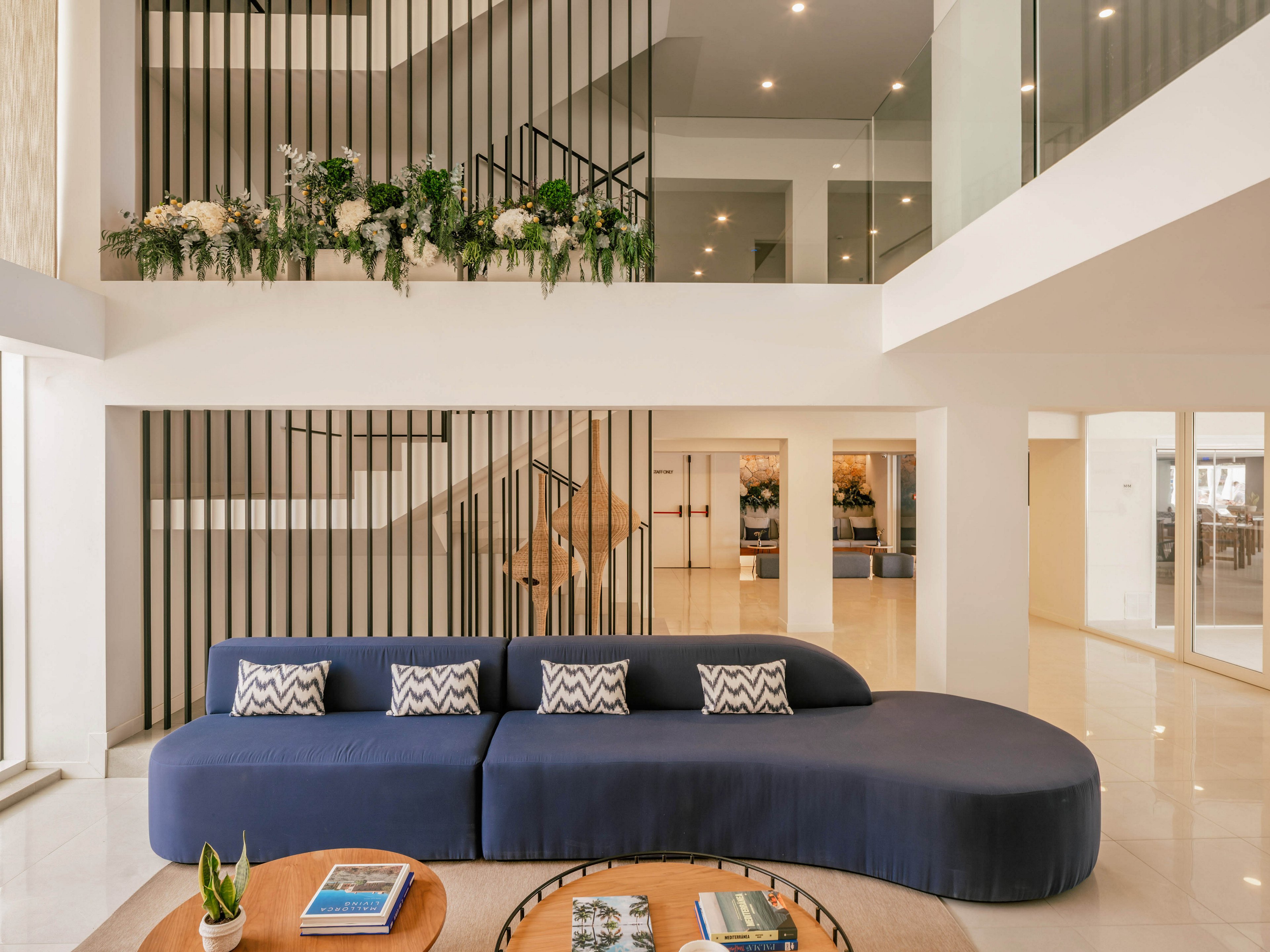 A contemporary lobby features a large curved navy blue sofa as the central focal point. The space is bright and open, with white walls, glossy floors, and multiple levels visible through glass railings. Vertical black metal slats create a striking backdrop, accented by green plants and white flowers. A round wooden coffee table in the foreground holds books and decorative items, enhancing the welcoming atmosphere.
