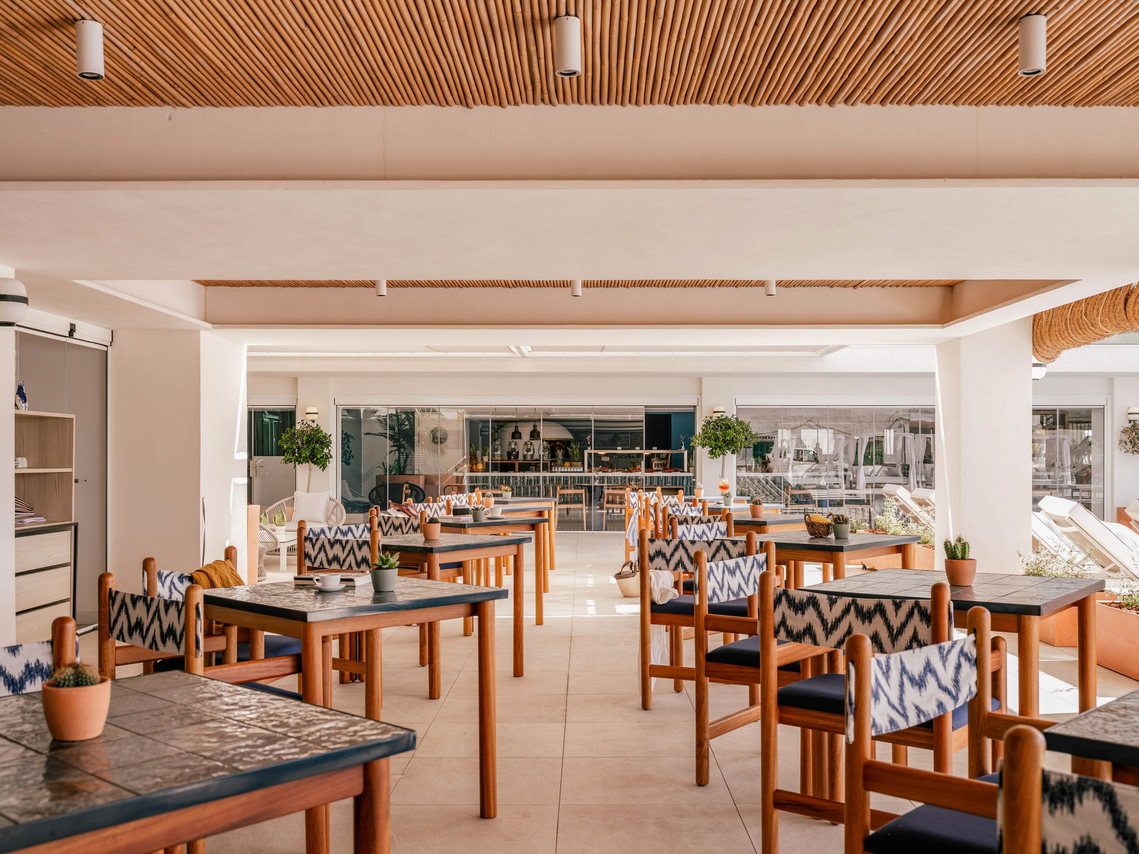 A contemporary open-air restaurant features neatly arranged wooden tables and chairs with blue and white patterned cushions. The space is bright and airy, with large openings leading to an outdoor area and a bar or service counter in the background. A warm wooden slat ceiling contrasts with the light walls and floor, creating a relaxed coastal atmosphere. Small potted plants on the tables add a touch of greenery and casual charm.