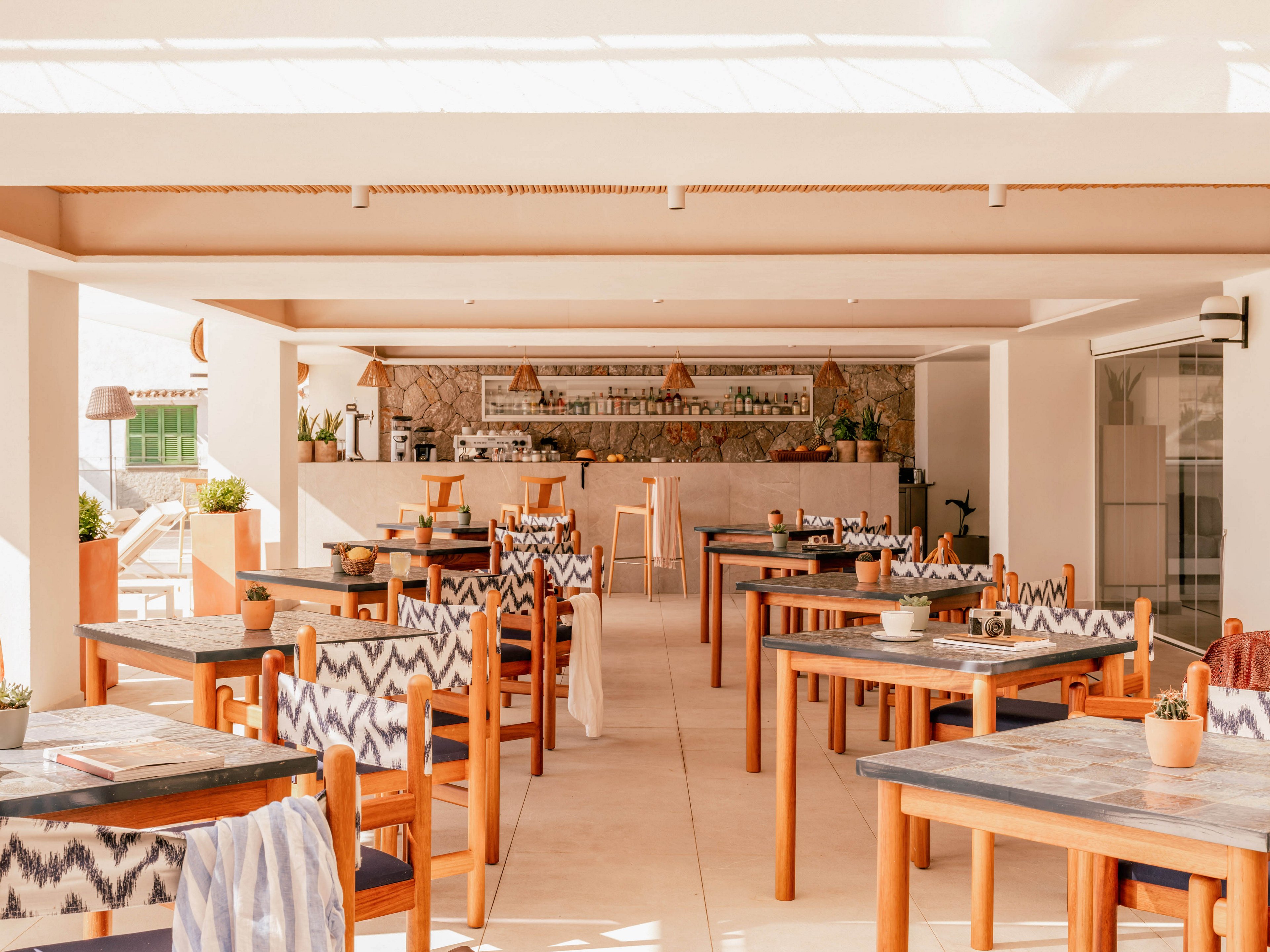 Sunlit wooden tables and chairs are arranged neatly in an open-air restaurant. The space features a central bar at the back with bottles and glassware displayed on shelves. Patterned chair backs and light neutral tones create a relaxed, coastal atmosphere. Potted plants and soft textiles add warmth and a casual, inviting feel to the dining area.