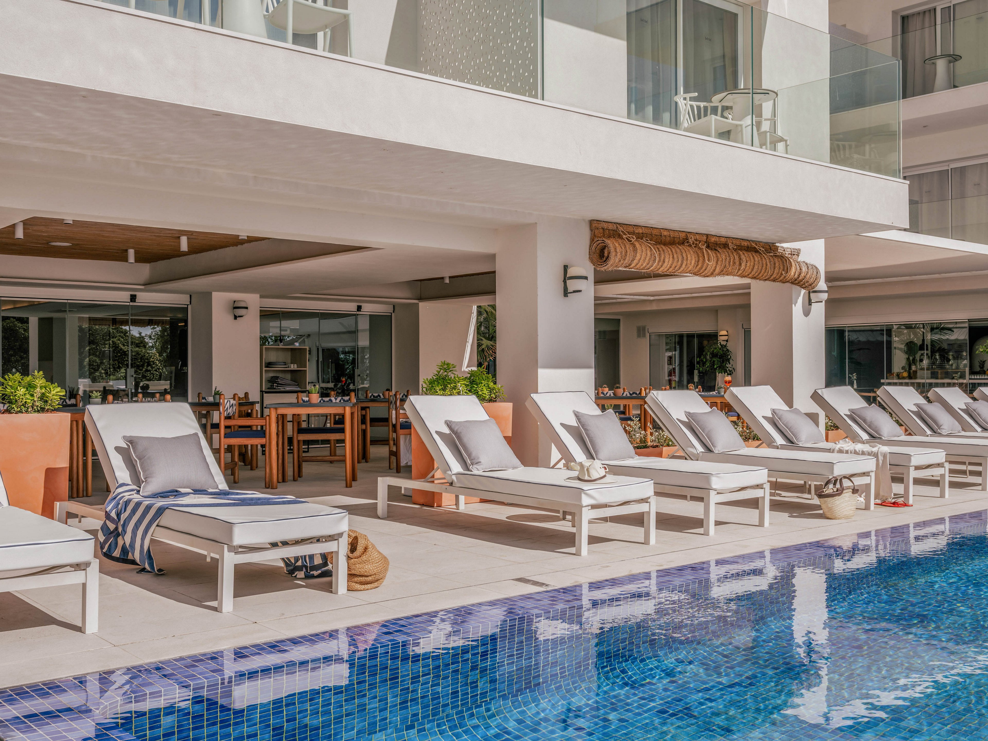 A row of white cushioned lounge chairs lines the edge of a clear blue tiled swimming pool at a contemporary resort. The scene includes striped towels, woven bags, and potted plants adding subtle decorative accents. Behind the loungers, an open-air dining area with wooden tables and chairs sits under a shaded overhang. Glass balcony railings and clean white architecture create a bright, minimalist atmosphere ideal for relaxation.