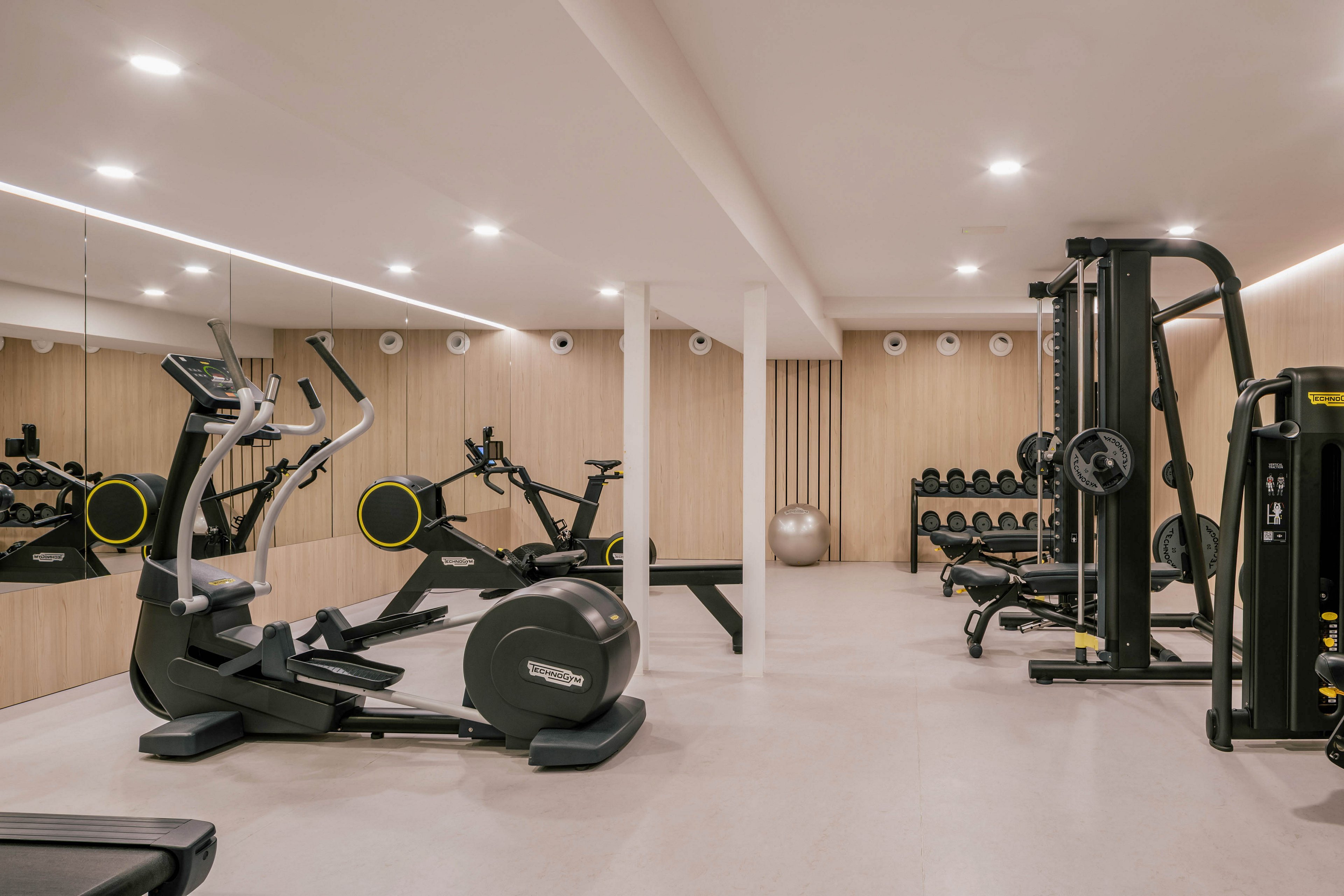 A contemporary home gym features various exercise machines arranged in a bright, minimalist room. Cardio equipment such as an exercise bike and treadmill sit near a mirrored wall, while strength machines and free weights line the opposite side. Light wood paneling and neutral flooring create a clean, calming atmosphere. Recessed ceiling lights evenly illuminate the space, highlighting the organized and uncluttered layout.
