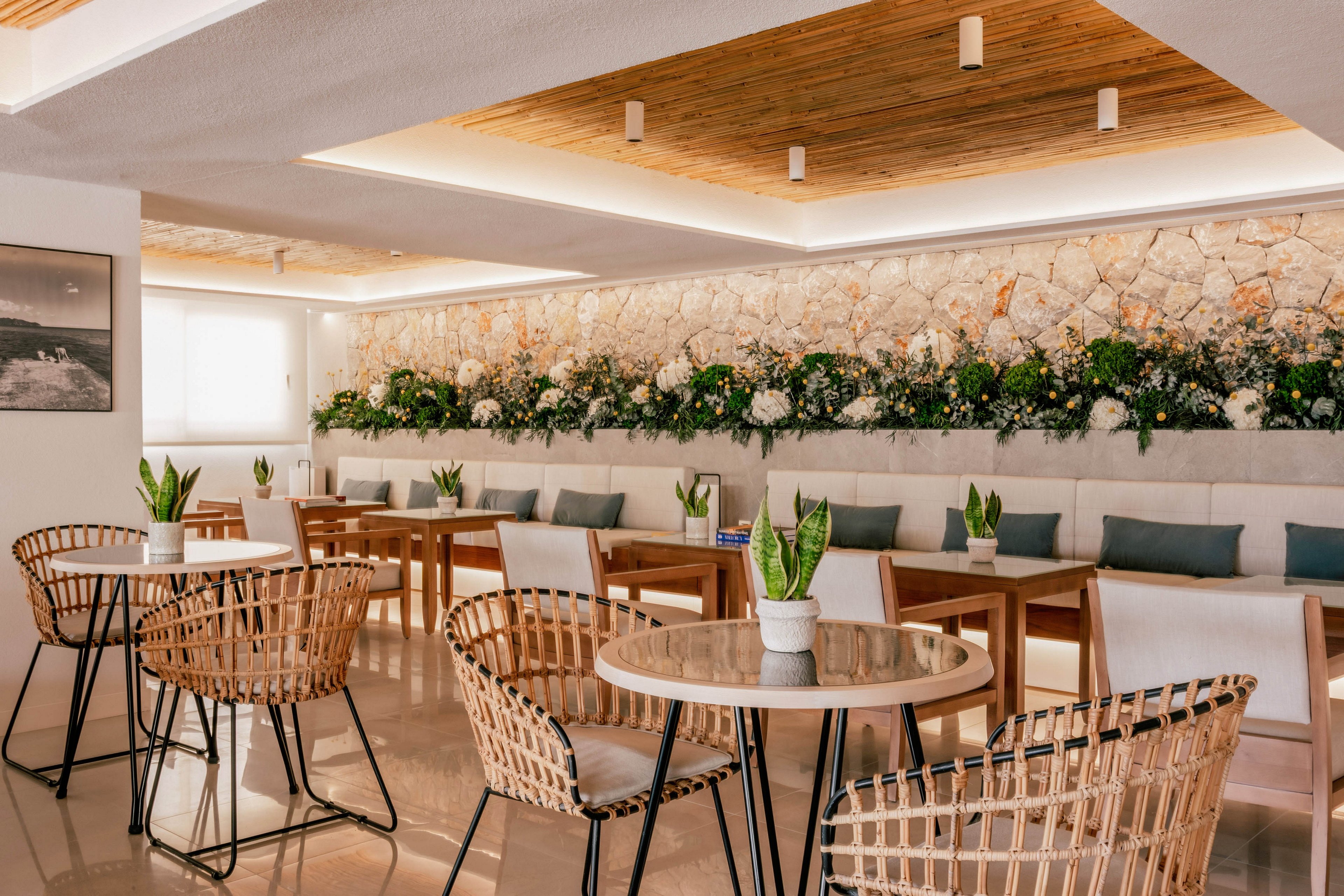 A bright, modern cafe interior features round tables with wicker chairs and small potted plants as centerpieces. Wooden tables and cushioned bench seating line the stone accent wall decorated with lush white and green floral arrangements. Warm recessed lighting in the wooden ceiling and reflective flooring create an inviting, contemporary atmosphere. The space appears calm and organized, ideal for relaxed dining or casual meetings.