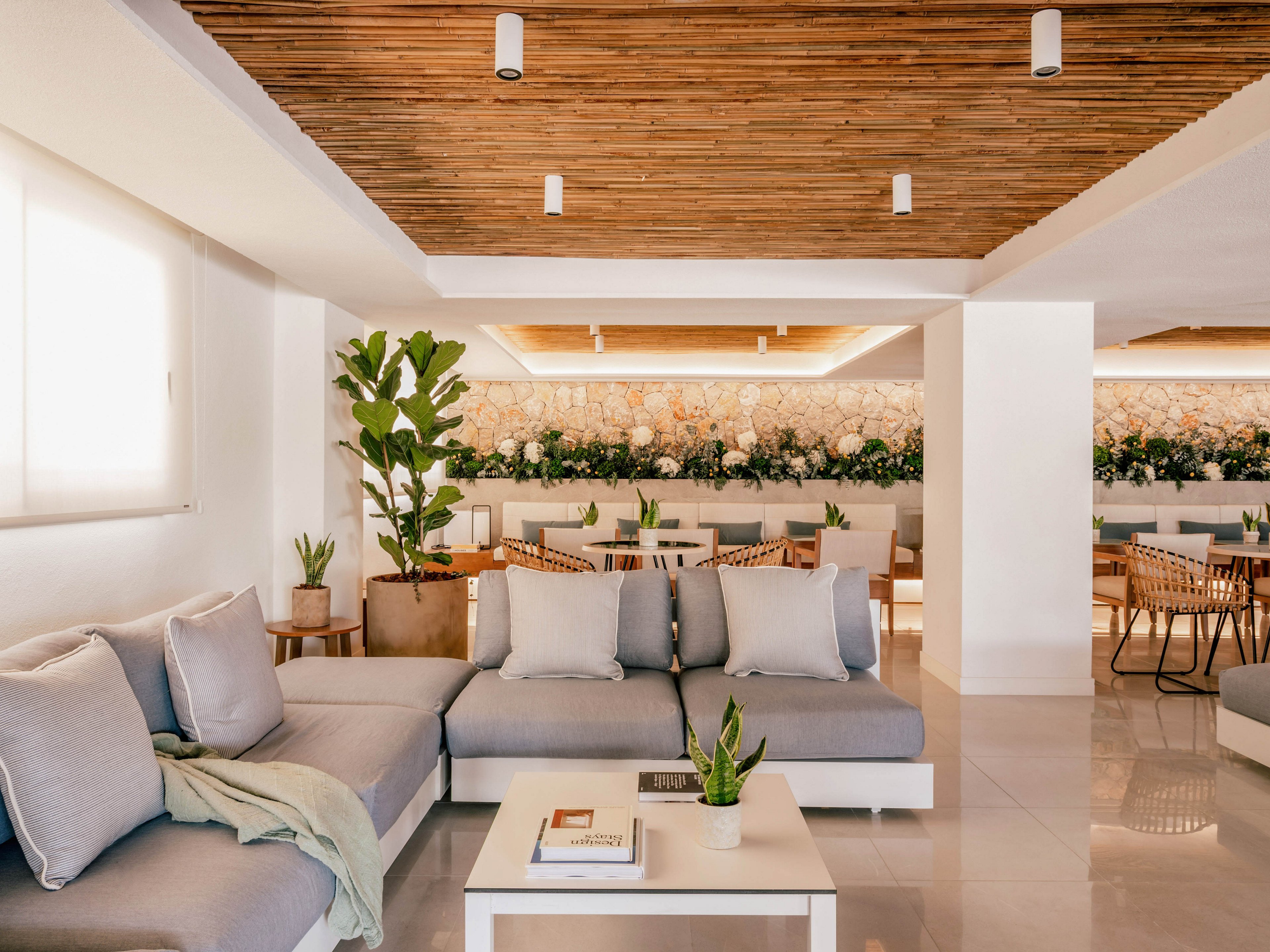A contemporary lounge area features light gray sofas with plush cushions and a small white coffee table. The space opens into a dining area with neatly arranged tables and woven chairs. Warm wooden ceiling panels and recessed lighting create a cozy yet sophisticated atmosphere. Potted plants and a long floral arrangement along the back wall add natural texture and color to the minimalist interior.