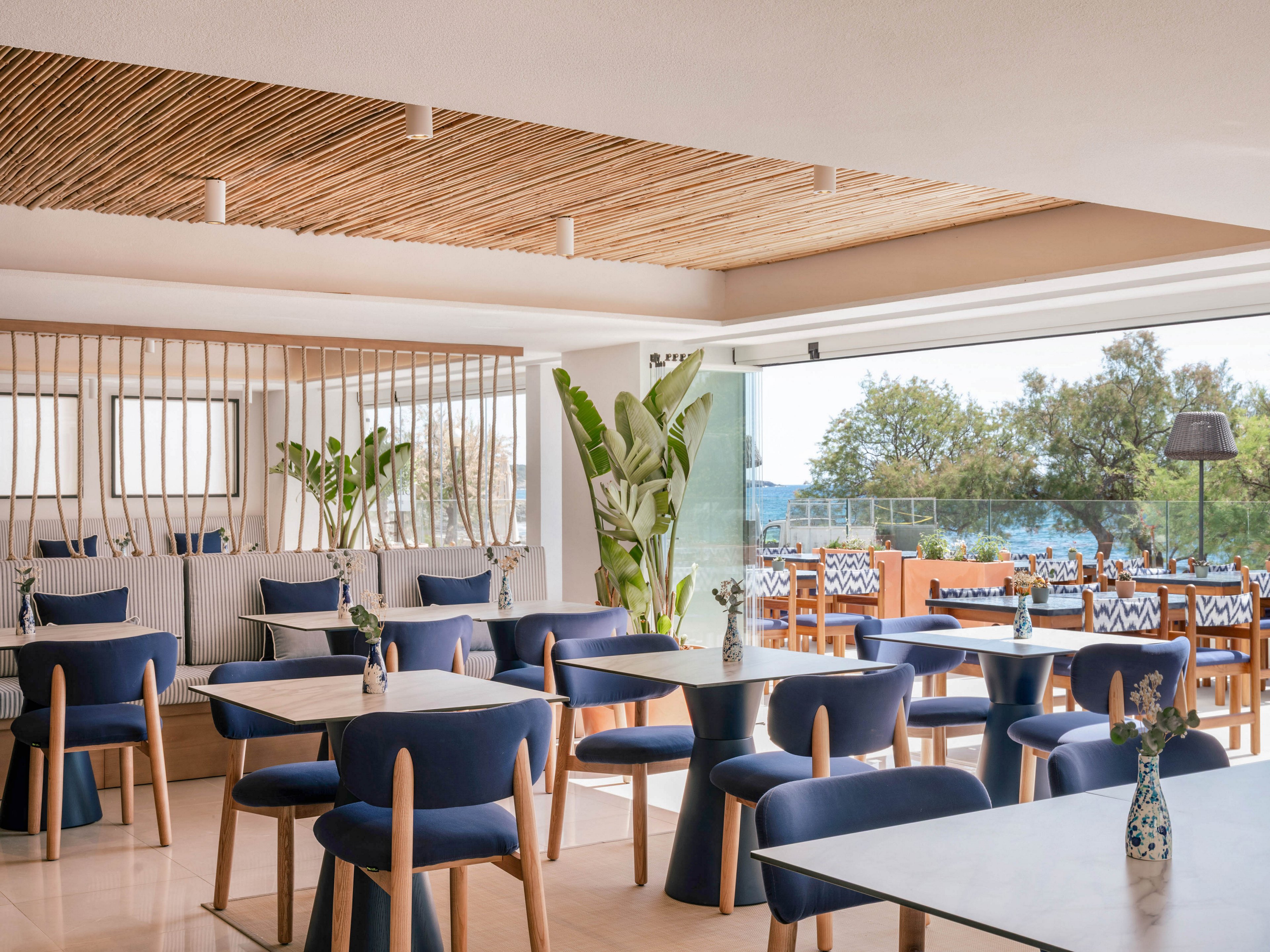 Contemporary restaurant interior features neatly arranged tables with blue cushioned chairs. Large open windows reveal an outdoor terrace dining area and a view of trees and water. Natural light fills the space, highlighting wooden ceiling slats, light flooring, and potted tropical plants. Decorative vases with flowers sit on the tables, creating a relaxed coastal atmosphere.