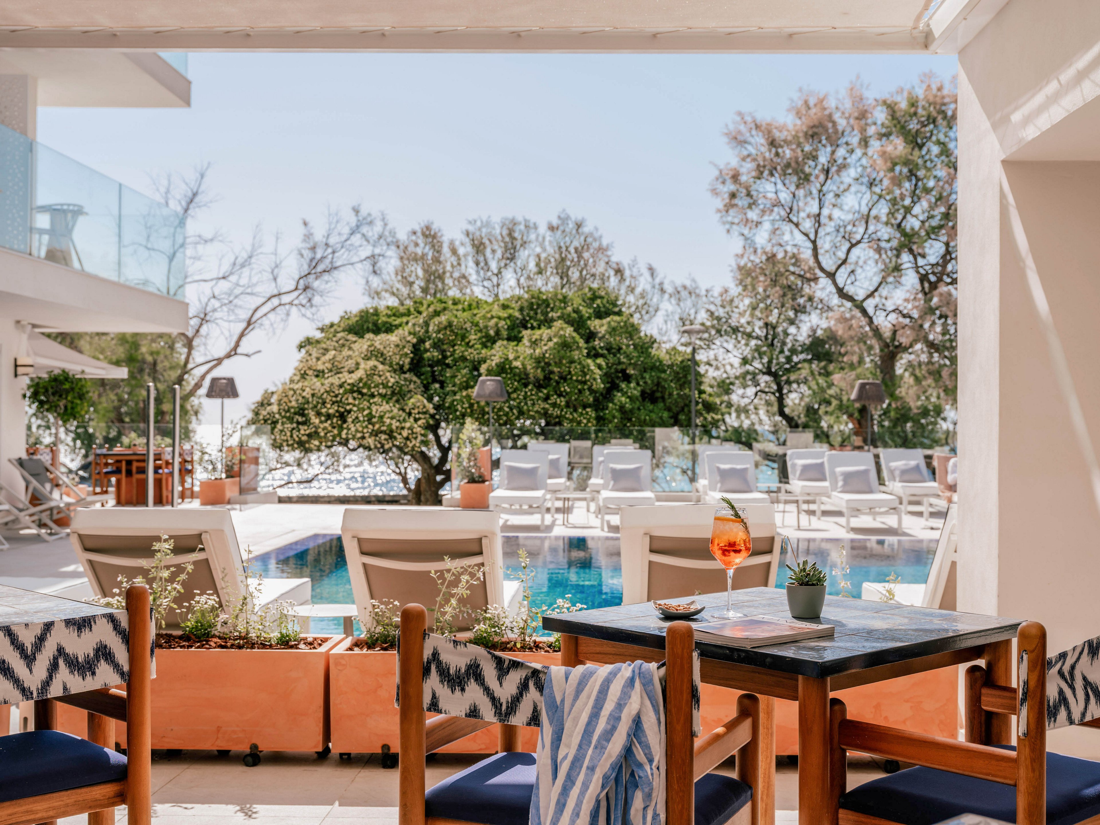 A stylish outdoor terrace overlooks a serene swimming pool lined with white sun loungers. In the foreground, a wooden table holds a bright orange cocktail and a small potted plant, with patterned cushions and a striped towel on the chairs. Trees and greenery frame the pool area, while soft natural light suggests a warm, sunny day. The scene conveys a relaxed vacation atmosphere at a modern resort.