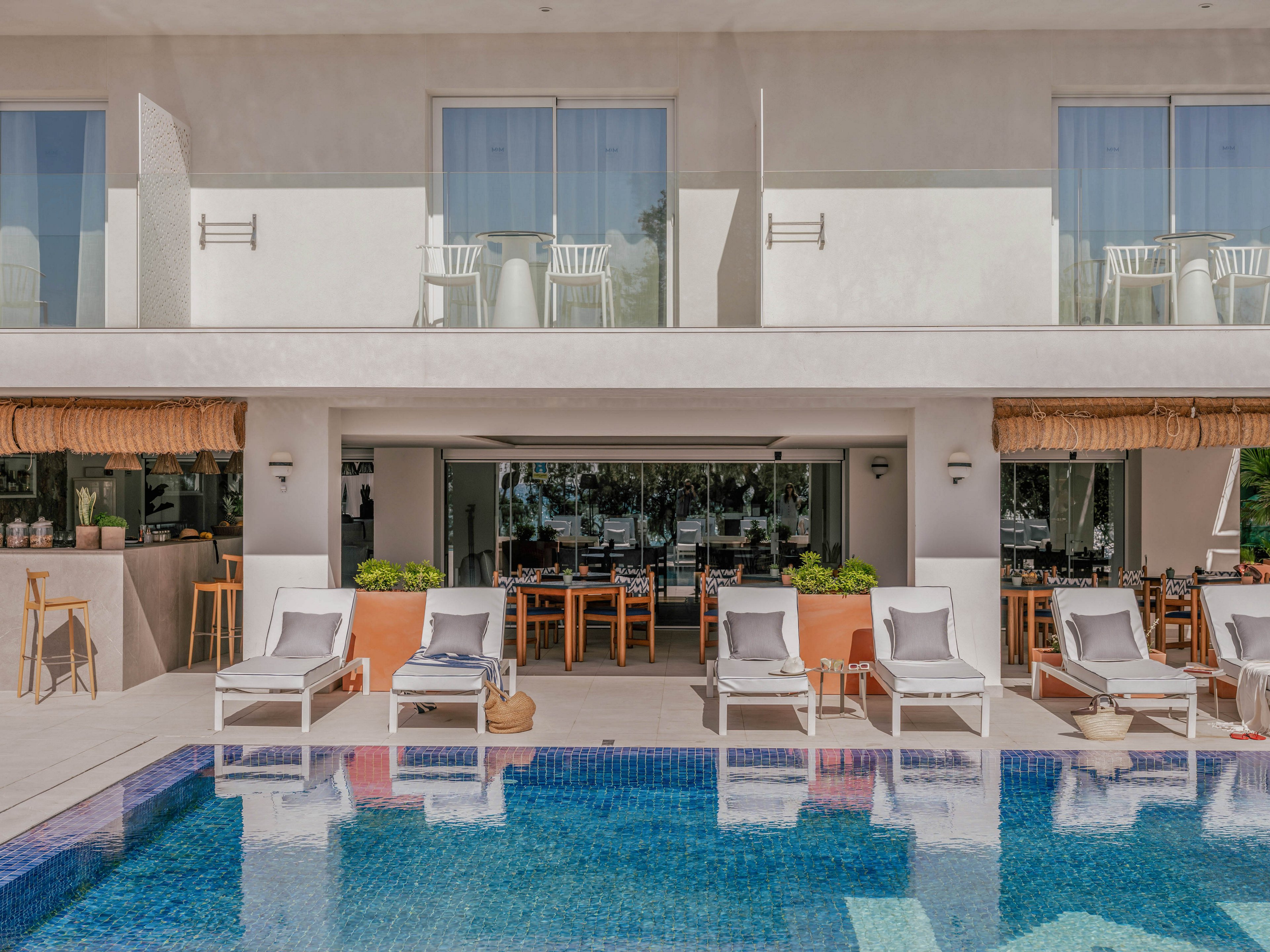 A contemporary resort pool area features white sun loungers lined up along a clear blue swimming pool. Behind the loungers, an open-air bar and dining area with wooden tables and chairs sits beneath a shaded terrace. Large potted plants add greenery to the neutral-toned architecture. Glass doors and upper-level balconies with sliding doors and chairs suggest guest rooms overlooking the pool. The overall scene feels calm, sunny, and inviting for vacation relaxation.