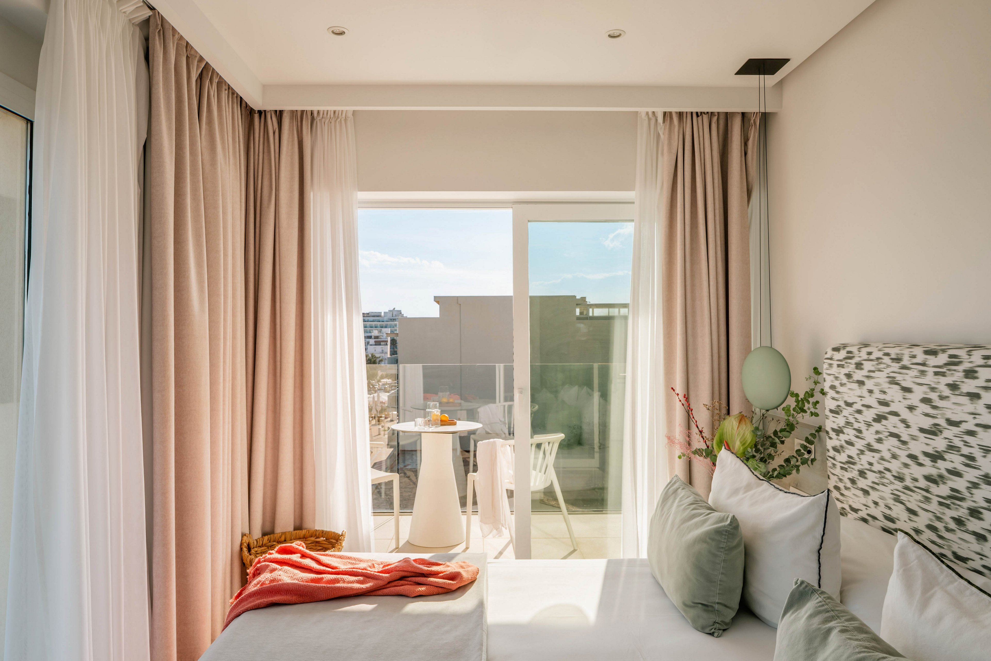 A sunlit modern bedroom features a neatly made bed with soft green and white pillows and a patterned headboard. Floor-to-ceiling curtains frame a large glass door that opens onto a small balcony. Outside, a round table with chairs and a tray of drinks or fruit is visible, suggesting a relaxing outdoor space. Warm natural light and pastel tones create a calm, inviting atmosphere ideal for rest and leisure.