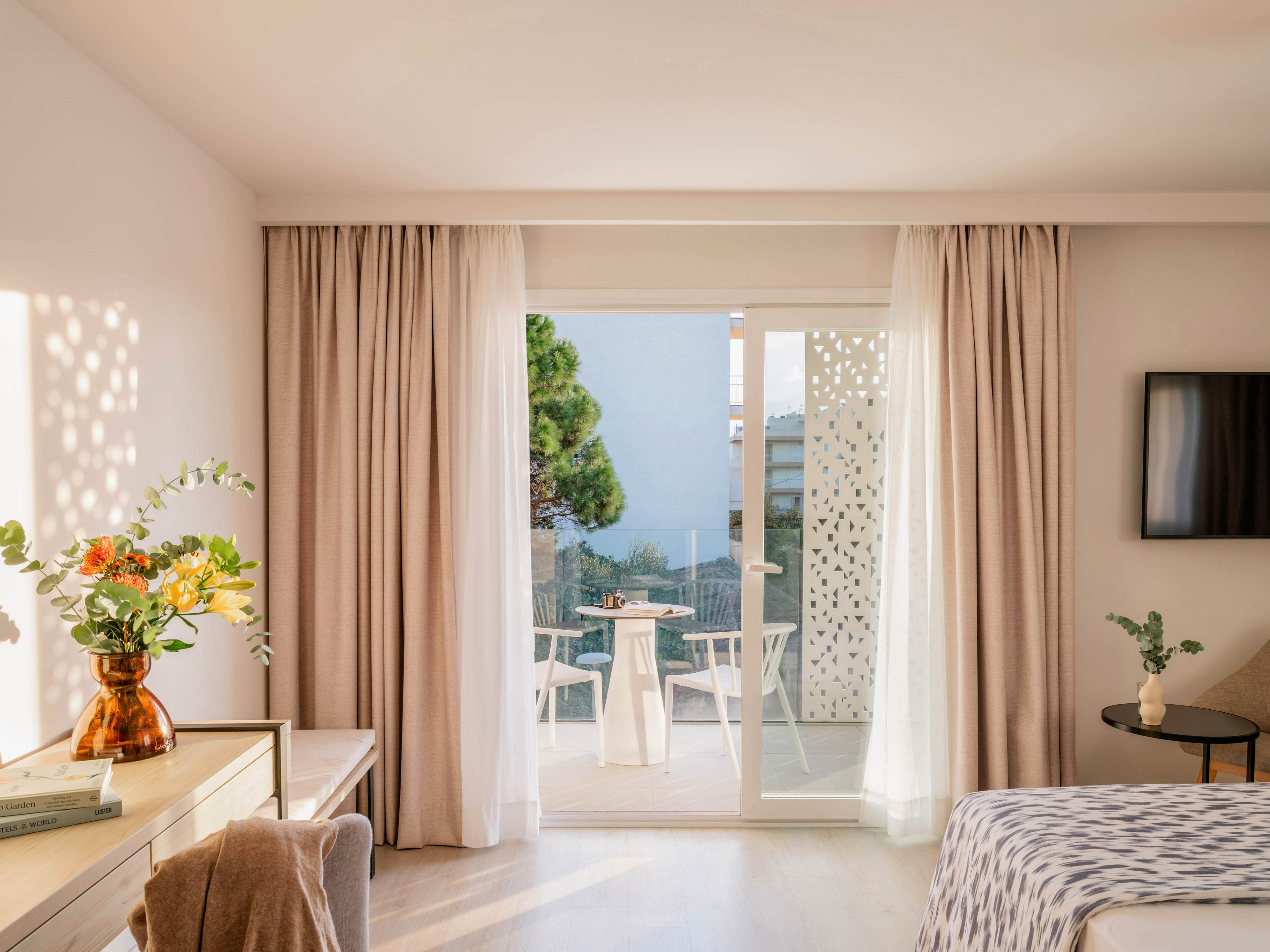 Bright, modern hotel room interior with a large glass door opening onto a balcony. Soft beige curtains frame the doorway, revealing an outdoor table and chairs and a distant view of trees and water. Warm sunlight fills the space, highlighting a neatly made bed, a console with flowers in a glass vase, and a wall-mounted television. The overall atmosphere is calm, airy, and inviting, ideal for a relaxing vacation stay.