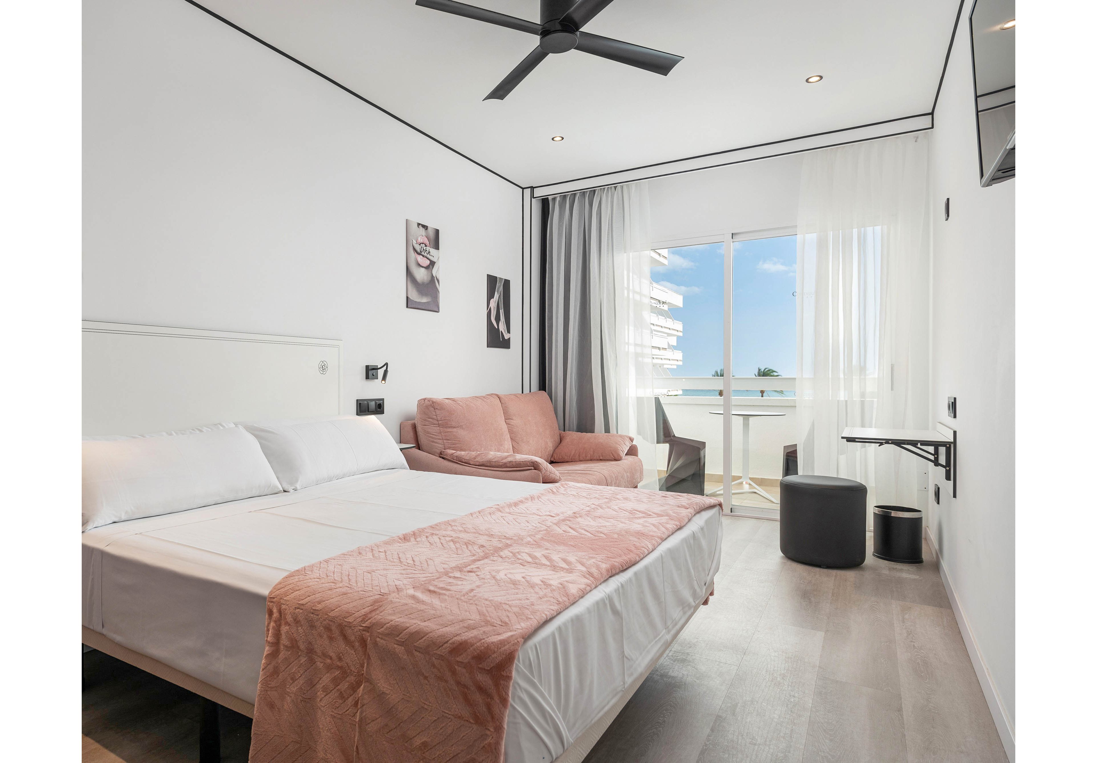 A contemporary hotel room features a double bed with white linens and a soft pink throw, a cozy seating area with a blush sofa, and a wall-mounted TV. The room is bright and airy, with large sliding glass doors opening to a balcony overlooking the ocean and nearby buildings. Minimalist decor, neutral tones, and natural light create a relaxing atmosphere, complemented by abstract wall art and sleek furnishings.