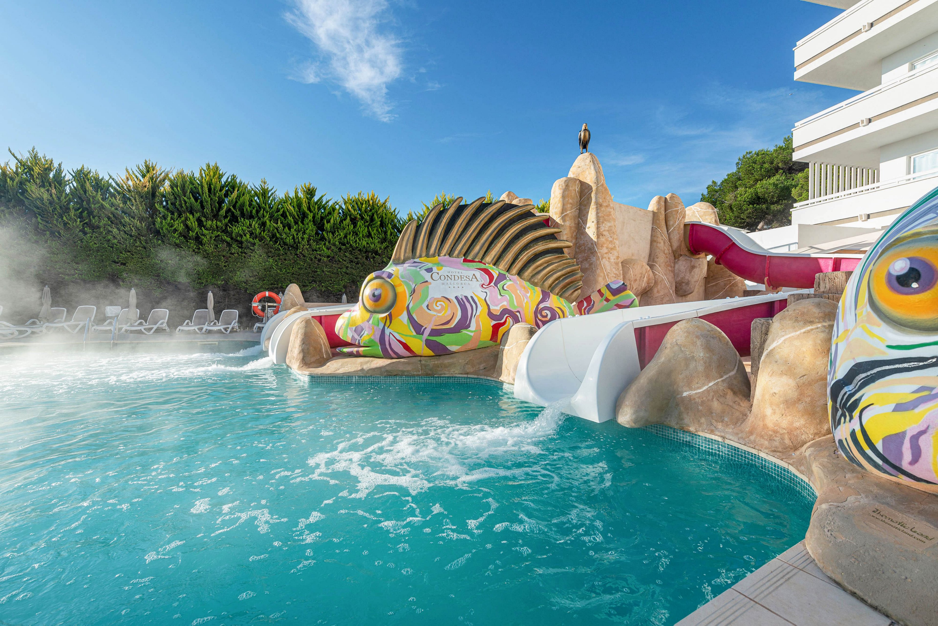 The main subject is a vibrant, fish-shaped water slide structure at an outdoor swimming pool. The setting features clear blue water, white lounge chairs, and lush greenery in the background, with a modern building visible to the right. The visuals include playful, multicolored designs on the slide and mist rising from the pool, creating a lively and inviting atmosphere. No visible text or numeric values are present in the image.