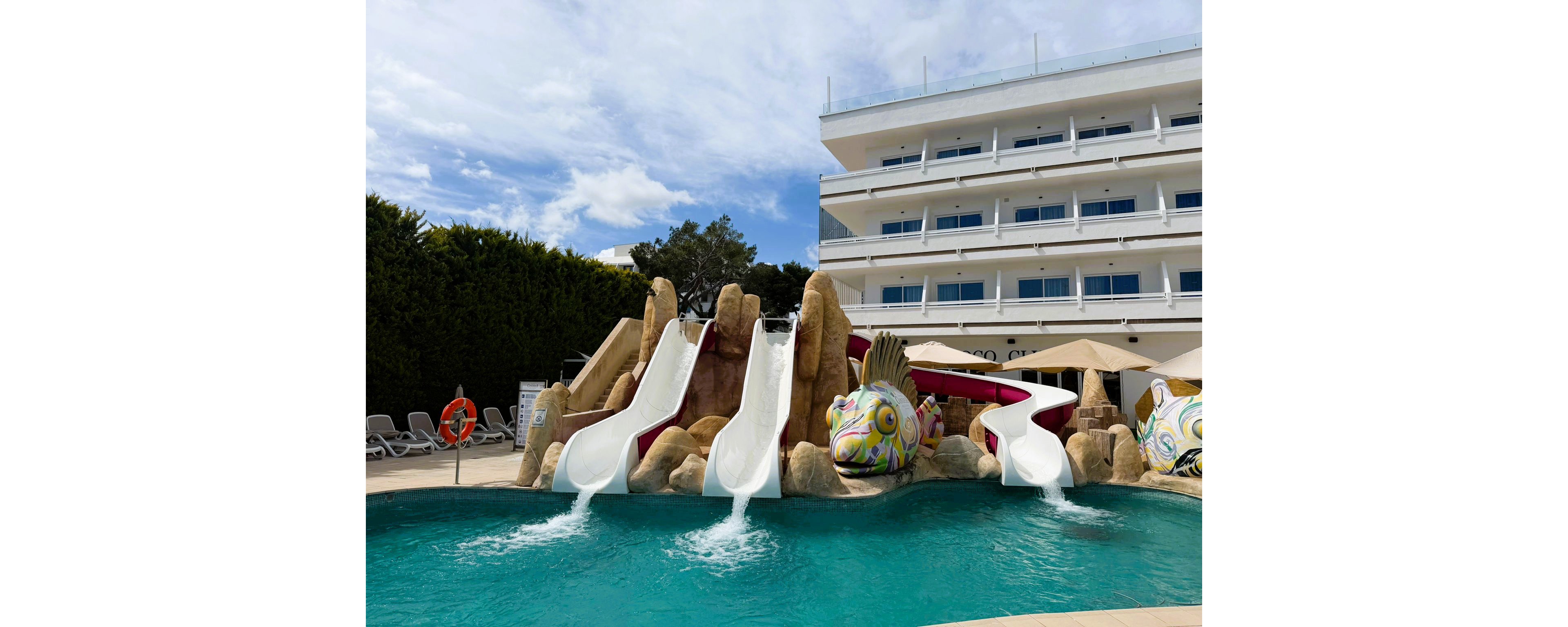 The main subject is a vibrant outdoor swimming pool featuring three white water slides emerging from artificial rock formations. Decorative fish sculptures add a playful touch to the pool area, which is set in front of a modern multi-story hotel building. Lounge chairs and umbrellas are visible, suggesting a relaxing and family-friendly atmosphere. The sky is partly cloudy, enhancing the bright and inviting mood.