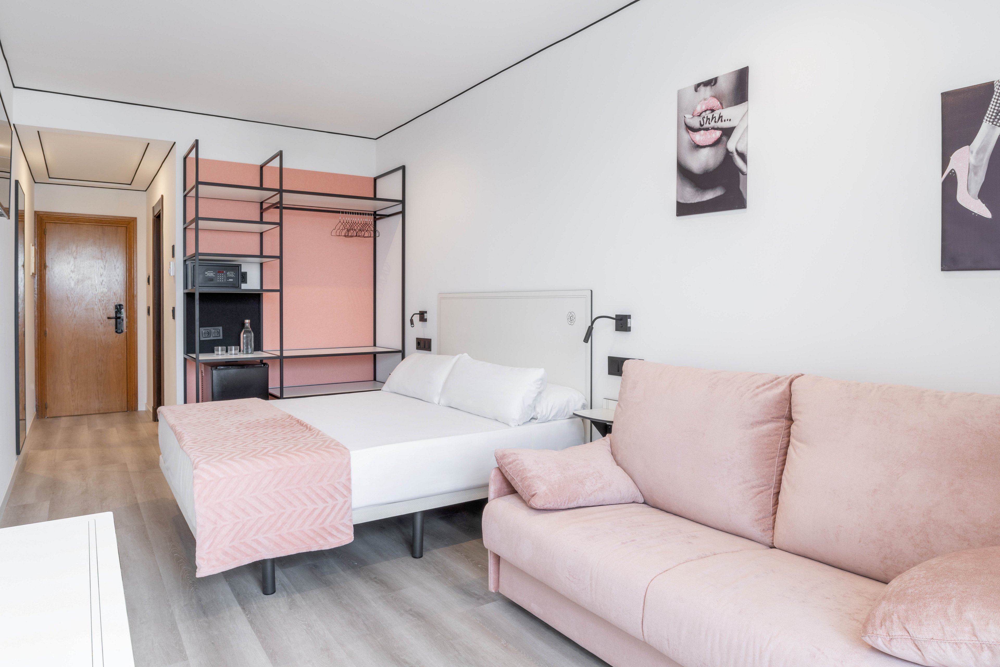 A contemporary hotel room features a double bed with white linens and a blush pink throw, complemented by a matching pink sofa. The space includes a built-in shelving unit with a microwave and safe, and wall art with close-up photography. The room is styled in a minimalist fashion with soft pastel tones and clean lines, creating a calm and inviting atmosphere.