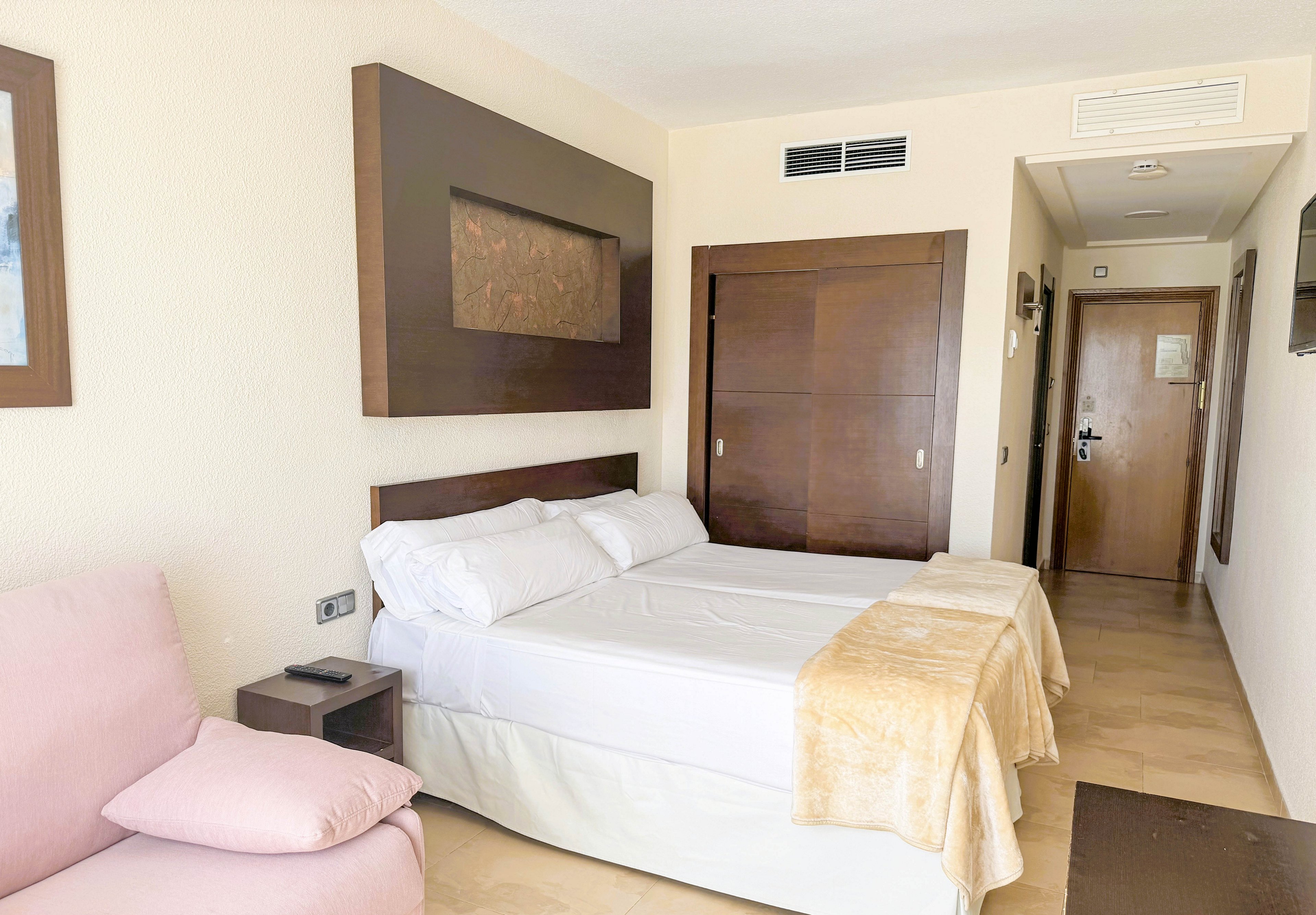 A contemporary hotel room features a double bed with white linens and beige throws, set against a cream-colored wall. The room includes dark wood accents, a small bedside table, and a light pink armchair. The hallway leads to a wooden door, and the overall palette is soft and neutral, creating a calm and inviting atmosphere.