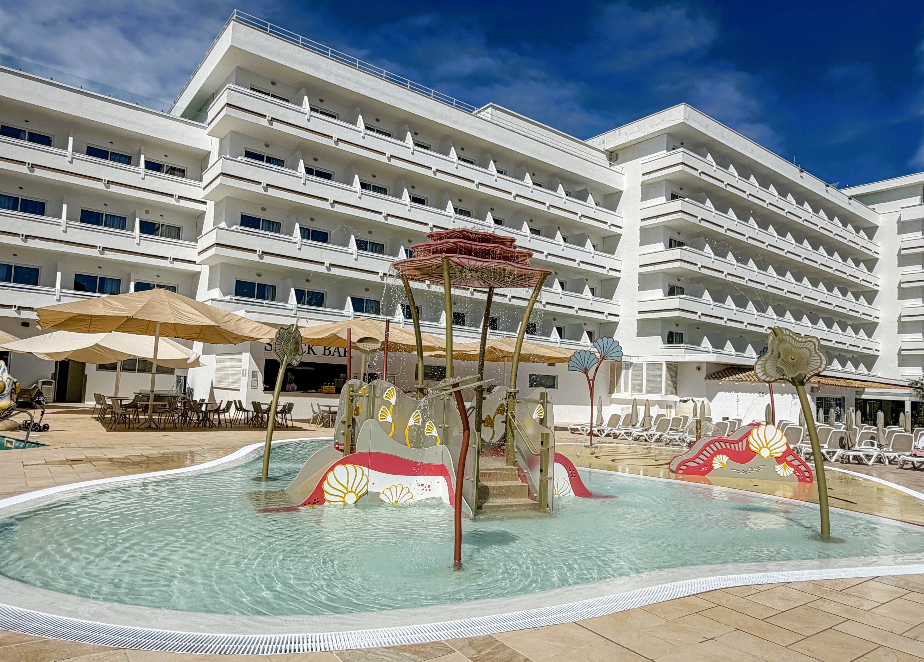 The image features a contemporary hotel building with multiple balconies and a spacious outdoor area. In the foreground, there is a shallow children's splash pool with whimsical water play structures and slides. Large umbrellas and lounge chairs surround the pool, creating a relaxed and family-friendly atmosphere. The sky is clear and blue, enhancing the bright and inviting mood.