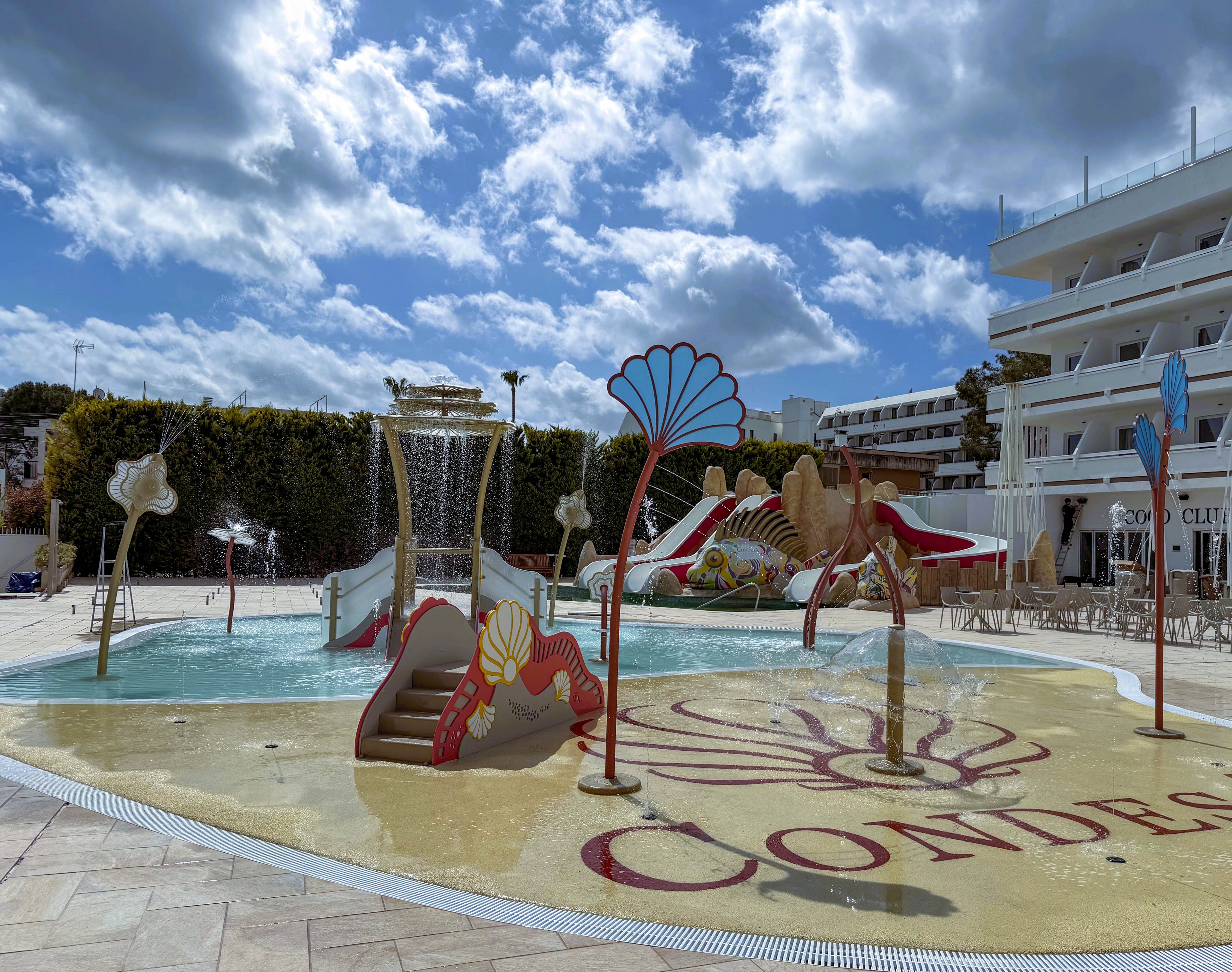 A colorful outdoor splash pool features playful aquatic-themed structures, including slides and water sprayers shaped like seashells and sea creatures. The pool is set in a resort environment with white buildings and lounge chairs visible in the background. The word 'CONDES' is clearly marked on the pool deck, and the area is bathed in natural daylight under a partly cloudy sky. No people are present in the scene.