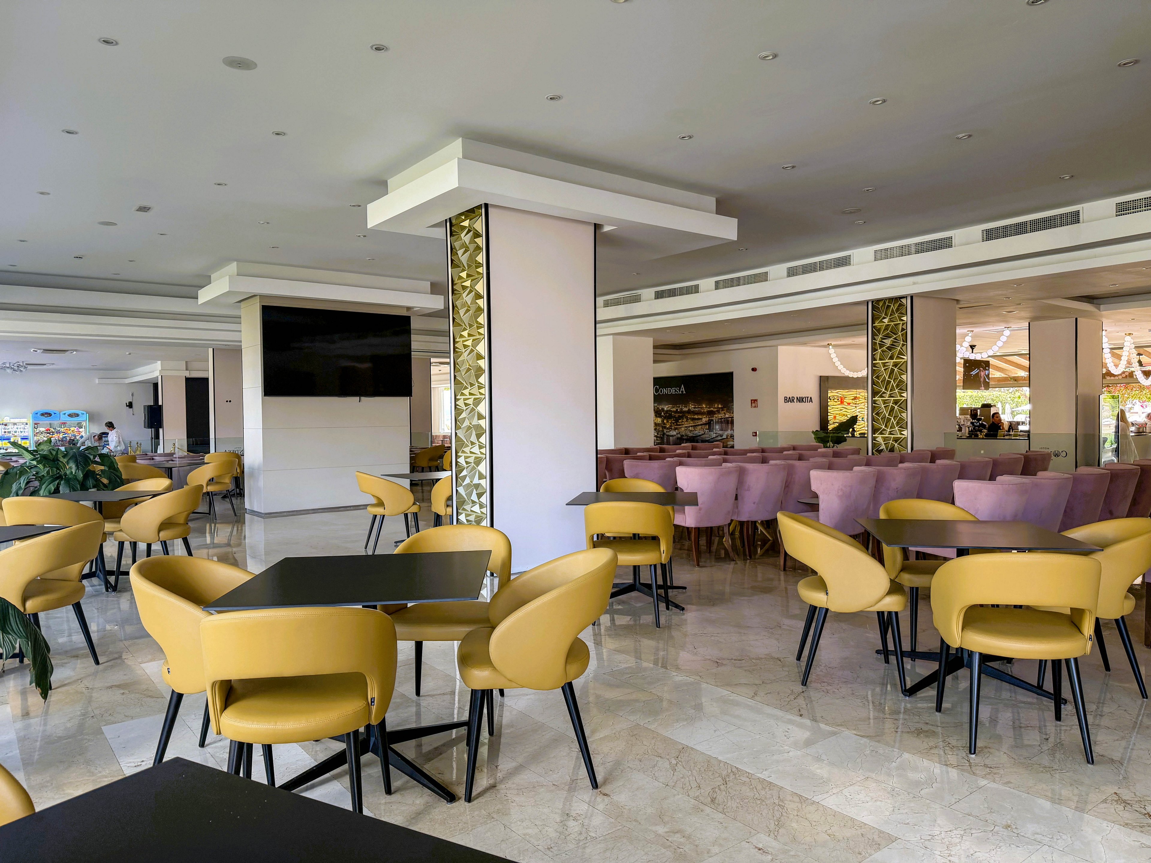 The image shows a spacious hotel restaurant featuring contemporary yellow chairs and black tables. The setting is bright with natural light, marble flooring, and decorative columns. The area is empty, highlighting the clean and organized layout, with purple seating visible in the background. No visible text or numeric values are present in the scene.