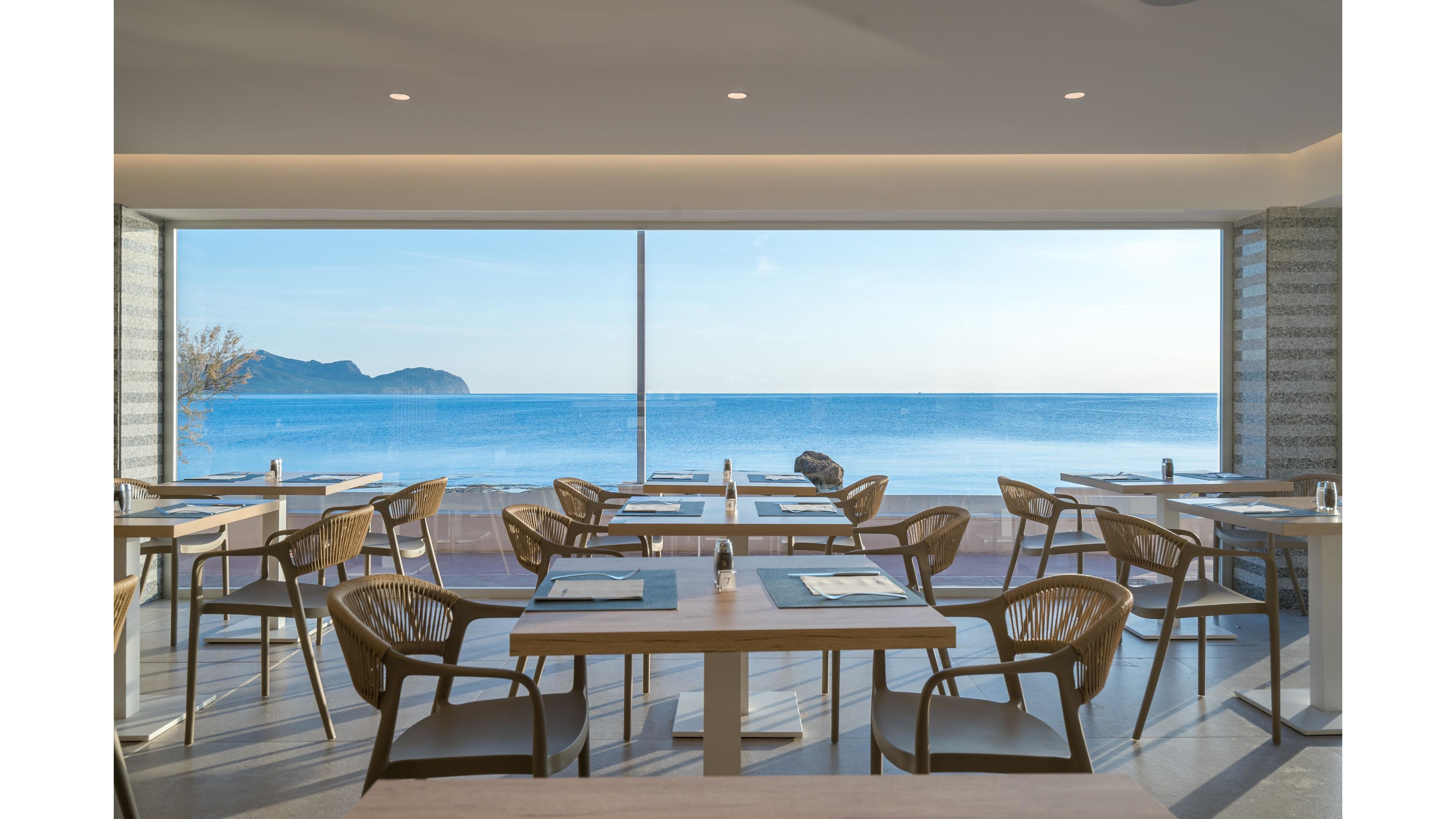 A contemporary restaurant dining area features neatly arranged wooden tables and woven chairs facing a large floor-to-ceiling window. Beyond the glass, a calm blue sea stretches to the horizon with a distant island and clear sky. Place settings with placemats and cutlery are prepared on each table, suggesting readiness for guests. The overall atmosphere is bright, minimalist, and tranquil, emphasizing the scenic coastal view.