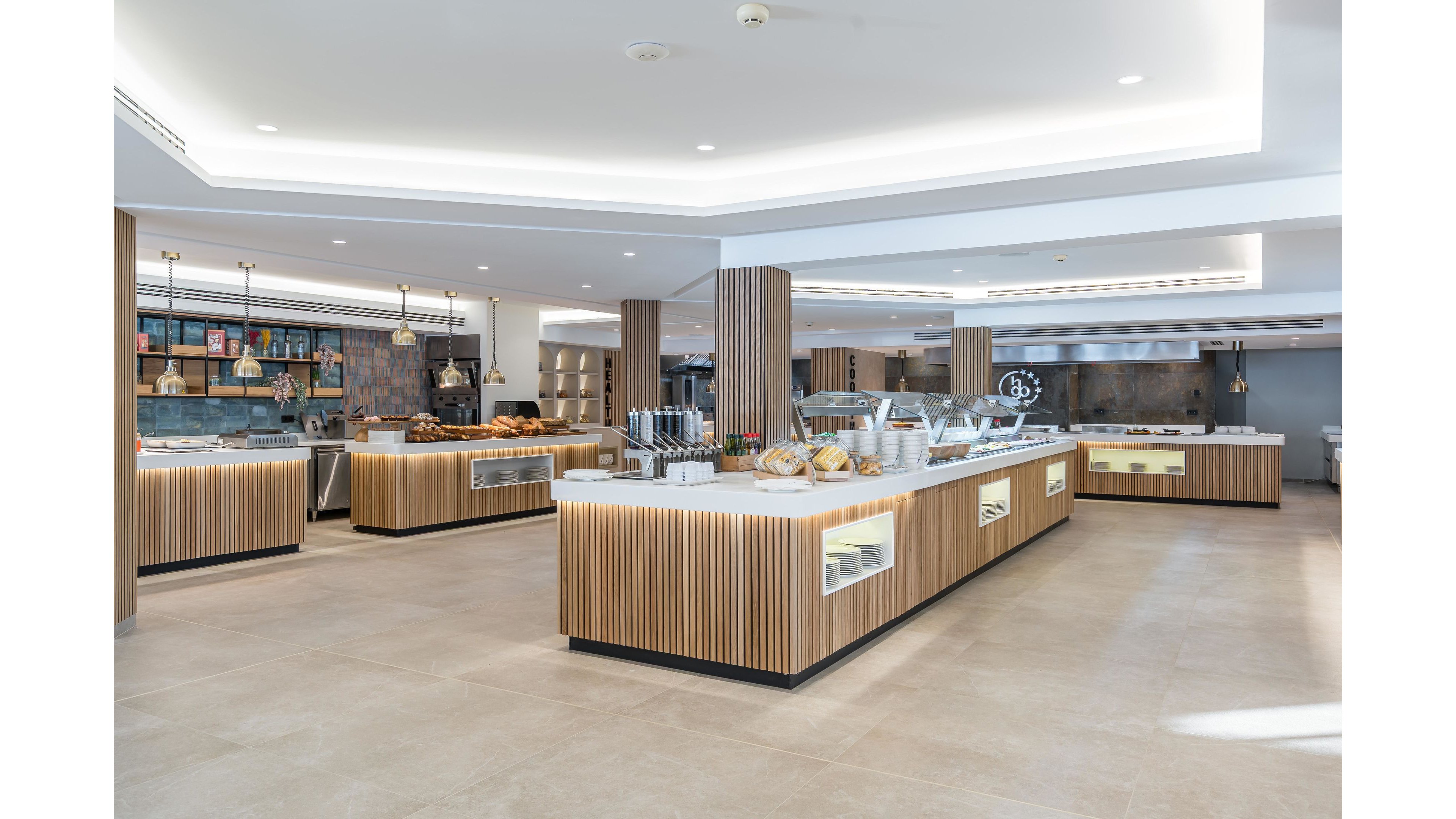 A spacious, modern buffet area features multiple wooden islands with white countertops arranged in an open-plan dining space. The counters display bread, pastries, and various food items under glass covers, suggesting a self-service setup. Warm wood paneling, recessed ceiling lighting, and neutral floor tiles create a clean, contemporary atmosphere. Shelving with jars and containers in the background adds visual interest and a casual, upscale feel.