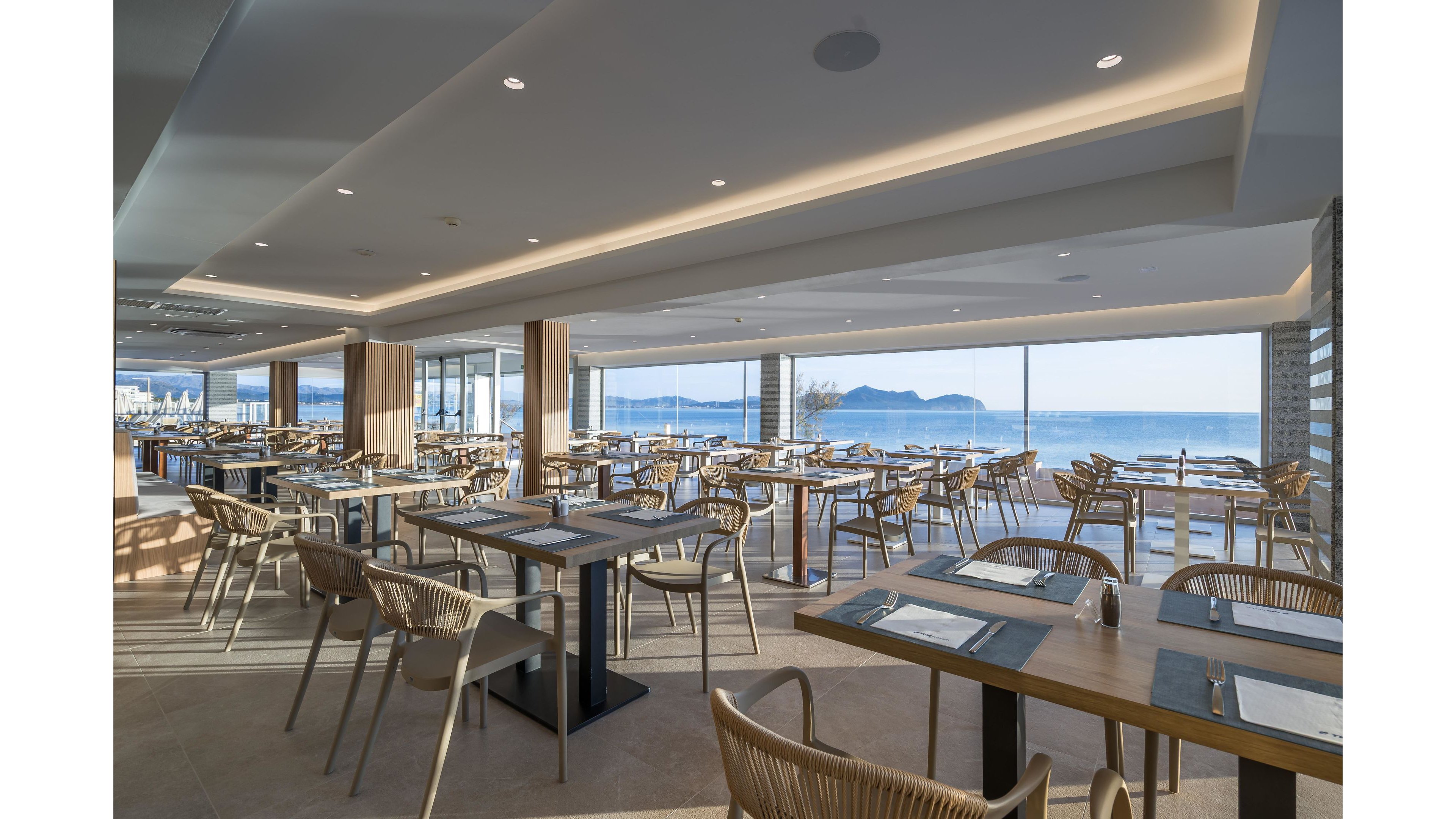 A spacious modern restaurant dining area features neatly arranged tables and wicker chairs. Large floor-to-ceiling windows reveal a bright seaside view with calm water and distant mountains. Neutral tones, clean lines, and soft ceiling lighting create a relaxed, contemporary atmosphere. The room appears ready for guests, with place settings laid out on each table.