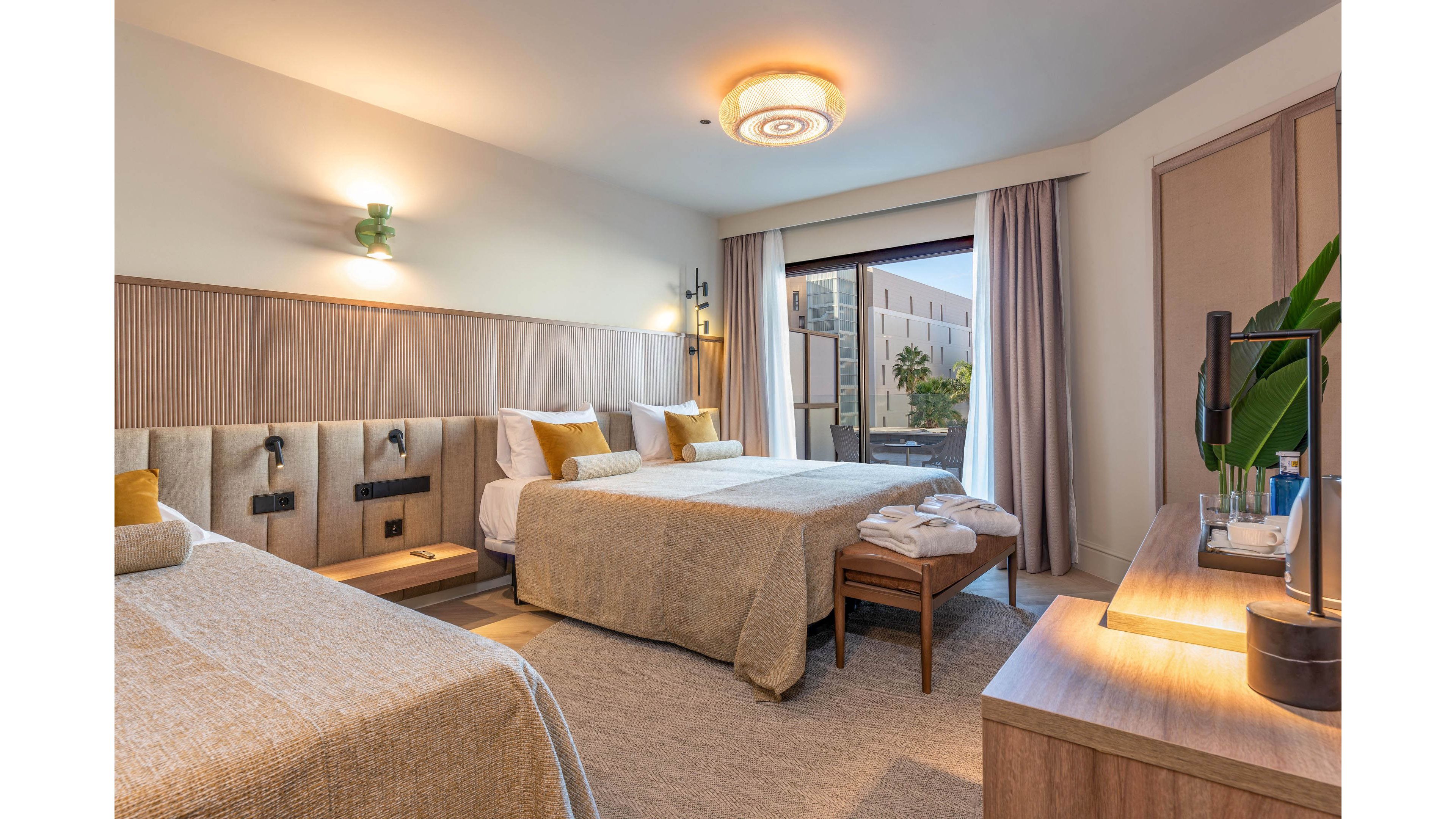 A contemporary hotel room features two large beds with neutral-toned bedding and mustard accent pillows. Warm wood furniture, including a bench and desk, complements the soft beige and cream color palette. A large window with sheer curtains opens to a balcony overlooking another building and palm trees. Soft ceiling and wall lighting create a cozy, inviting atmosphere suitable for relaxation or business travel.