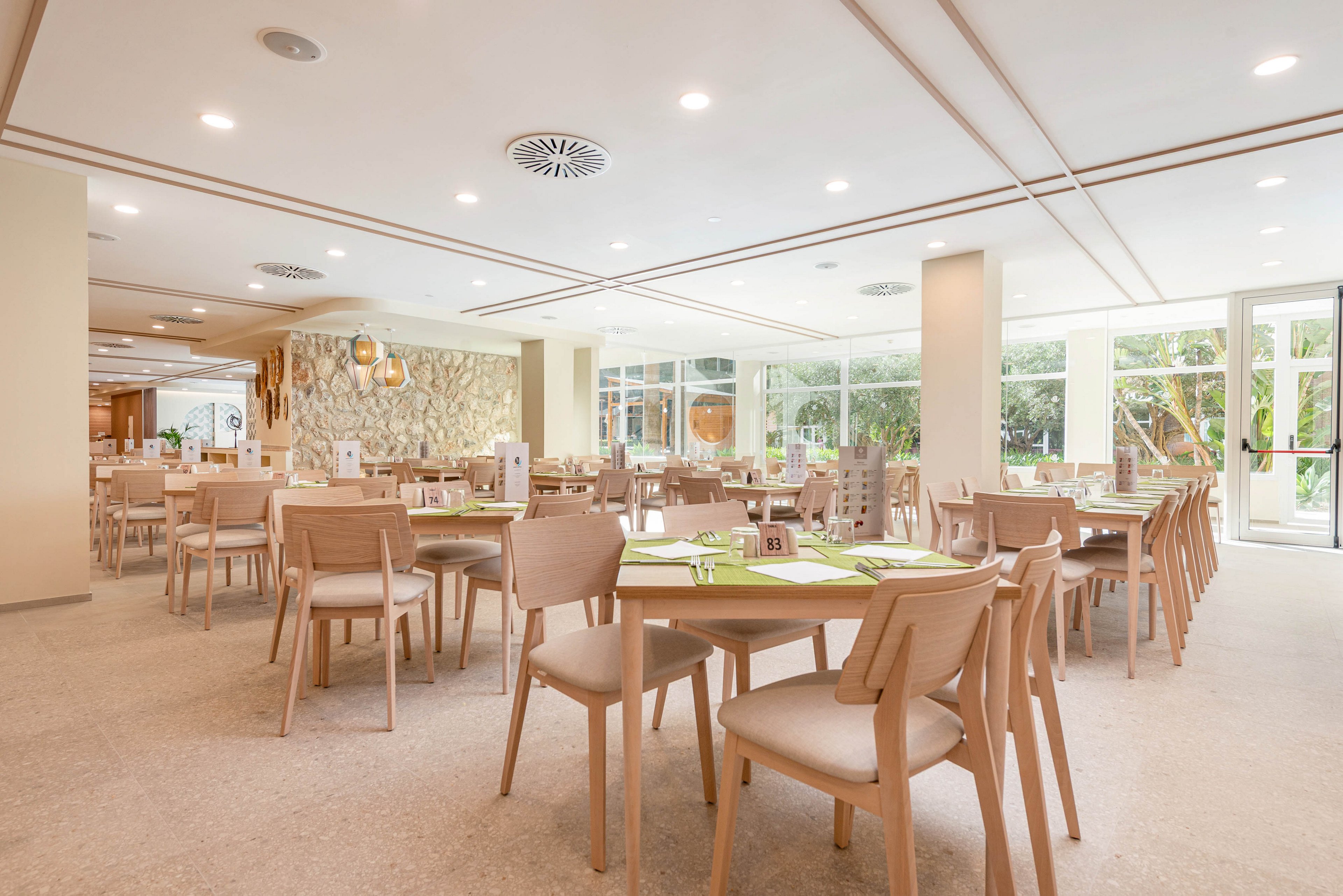 Spacious restaurant dining room with light wooden tables and chairs neatly arranged. The setting features large floor-to-ceiling windows that let in abundant natural light and offer views of greenery outside. Neutral tones dominate the space, complemented by green placemats and simple table settings. Ceiling lights and contemporary decor create a clean, modern, and welcoming atmosphere.