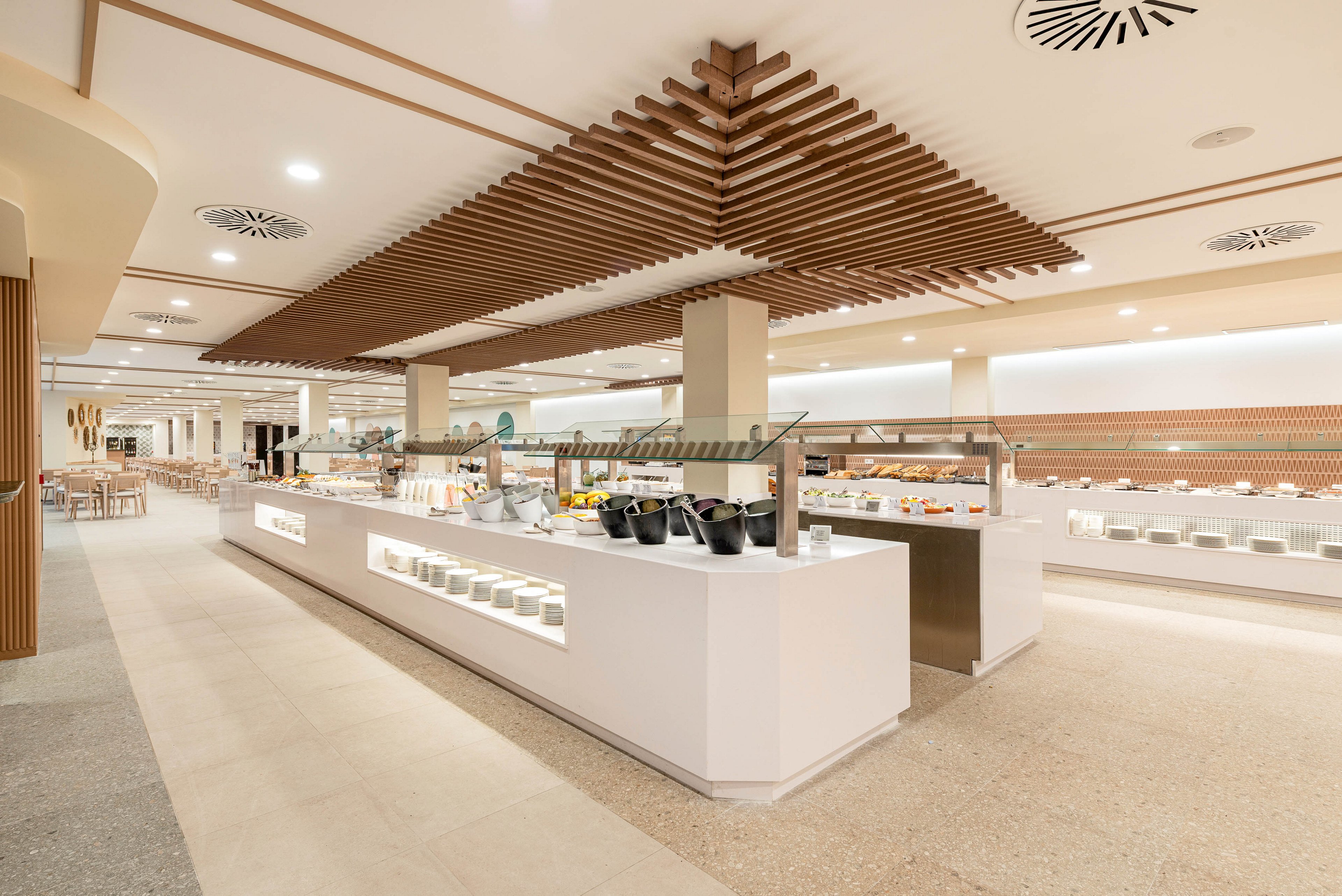 A spacious self-service buffet area is shown in a modern restaurant or hotel dining room. Long white counters display various dishes and utensils under bright, even lighting. Wooden ceiling panels and neutral-toned flooring create a clean, contemporary aesthetic. The seating area extends into the background, emphasizing the open-plan layout and minimalist design.