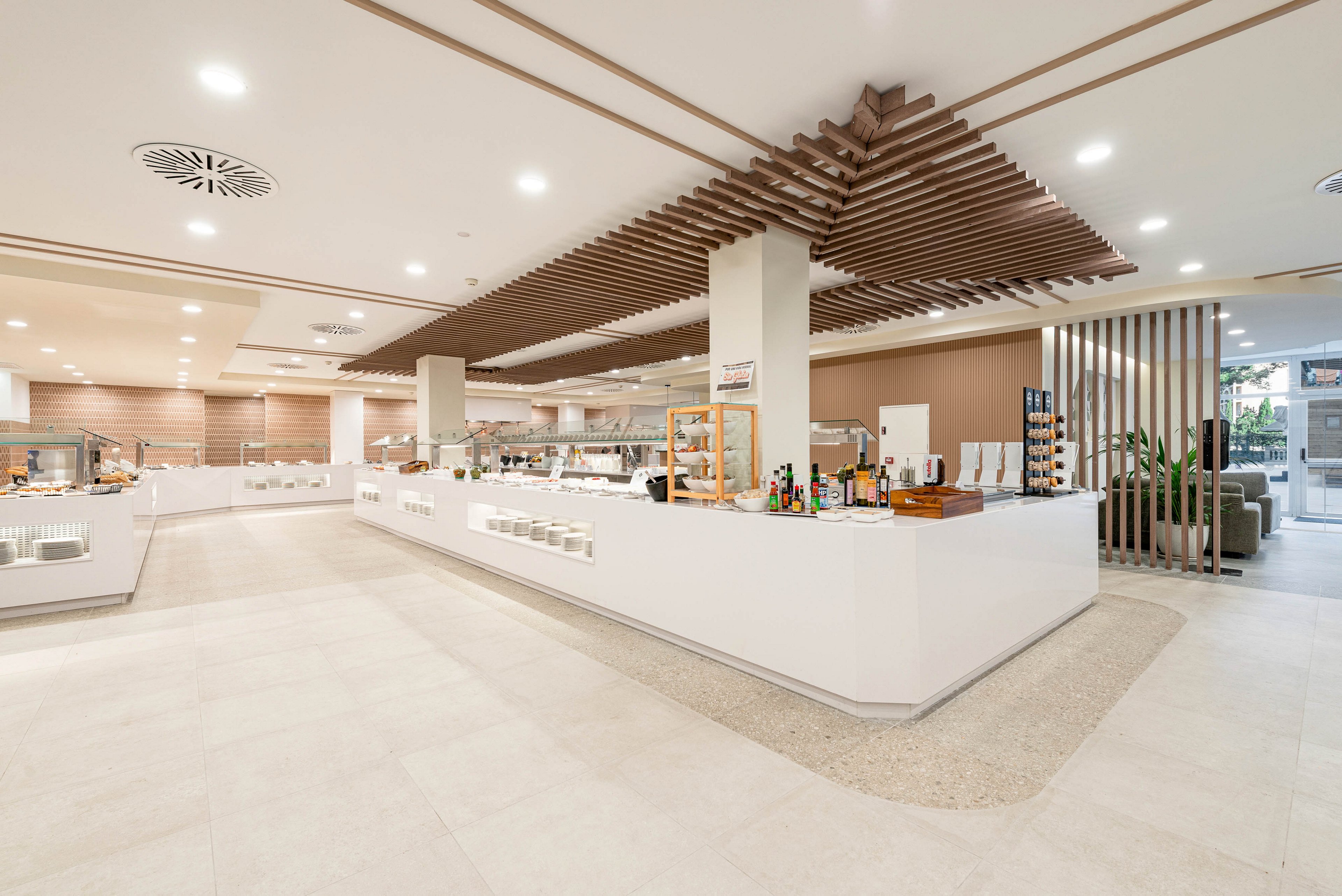 A spacious, modern buffet area features long white counters arranged around a central column. The setting appears to be a hotel or restaurant dining space prepared for self-service meals. Bottles, plates, and food displays are neatly organized along the counters, with warm neutral tones and clean lines throughout. Recessed lighting and decorative ceiling panels create a bright, contemporary atmosphere. The room is empty of people, emphasizing the open layout and minimalist design.