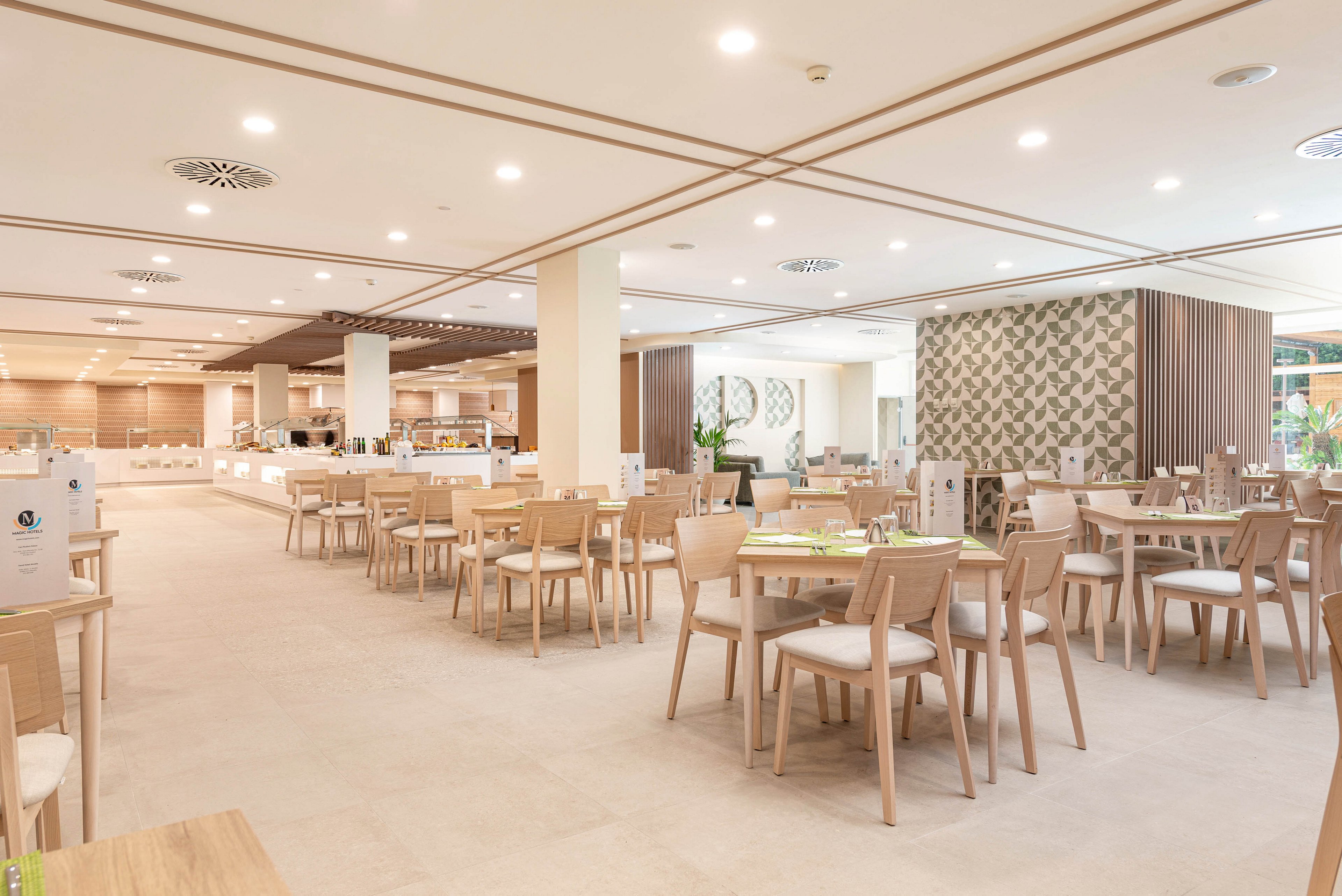 Spacious modern restaurant interior with light wooden tables and chairs arranged neatly. The setting appears to be a hotel or resort dining area with a buffet section visible in the background. Neutral beige tones, soft lighting, and clean lines create a calm and welcoming atmosphere. Decorative wall panels and ceiling details add a contemporary design touch to the open space.