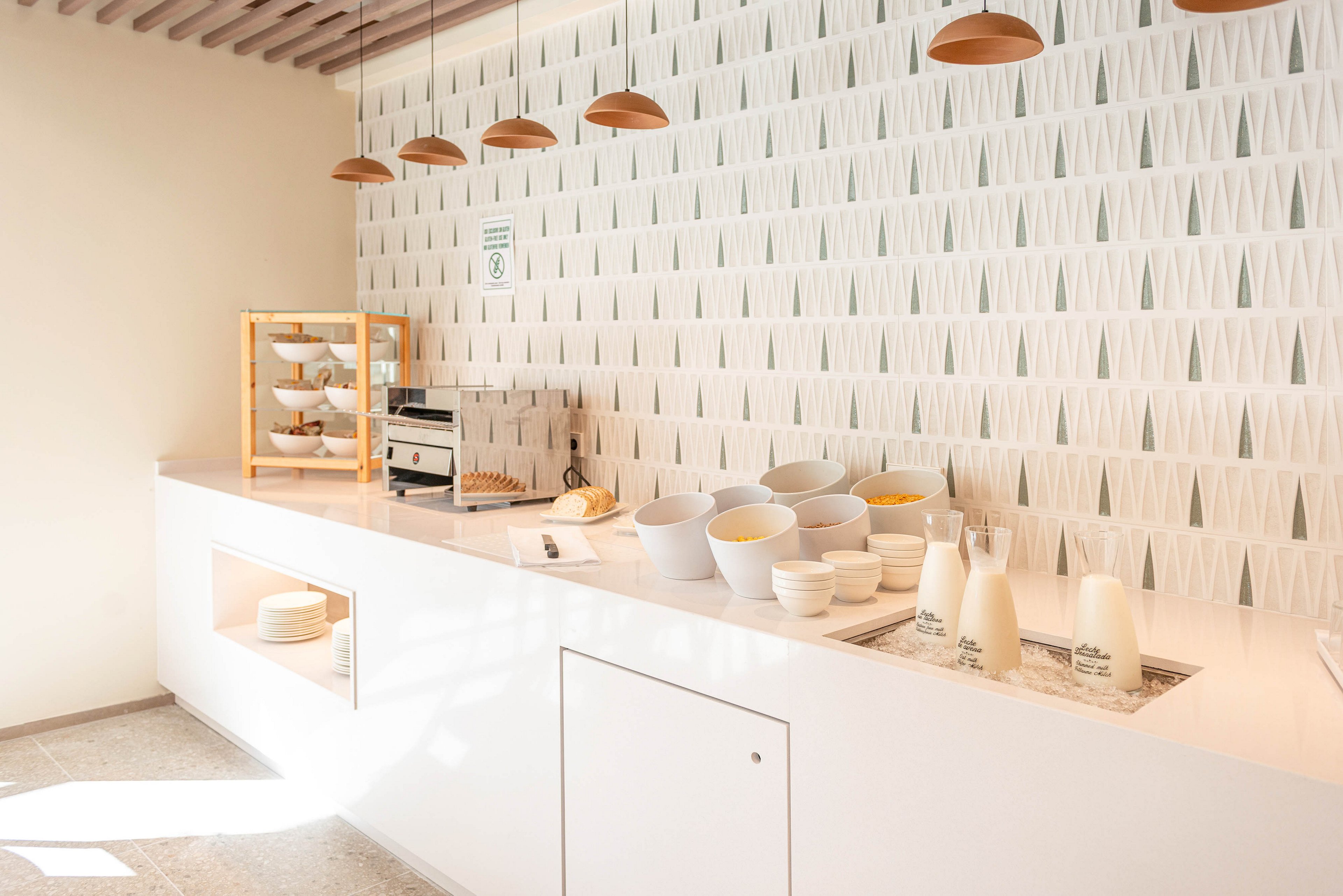 A bright, modern breakfast buffet counter is set in a minimalist interior. White bowls filled with cereal and glass bottles of milk are neatly arranged on the countertop beside stacks of plates. A wooden display case holds pastries near a stainless steel toaster. Warm pendant lights hang from the ceiling, complementing the geometric patterned wall in soft neutral tones. The overall scene feels clean, organized, and inviting.