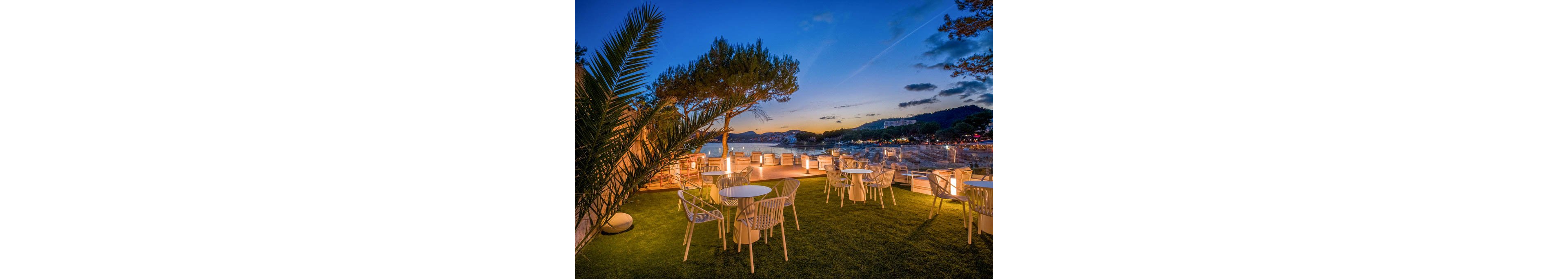 An outdoor terrace lounge overlooks a calm coastal bay at sunset. White chairs and round tables are arranged on a green lawn, facing the water and softly glowing lights along the railing. The sky transitions from deep blue to warm orange tones as the sun sets behind distant hills. Palm fronds and trees frame the scene, creating a relaxed, upscale seaside atmosphere.