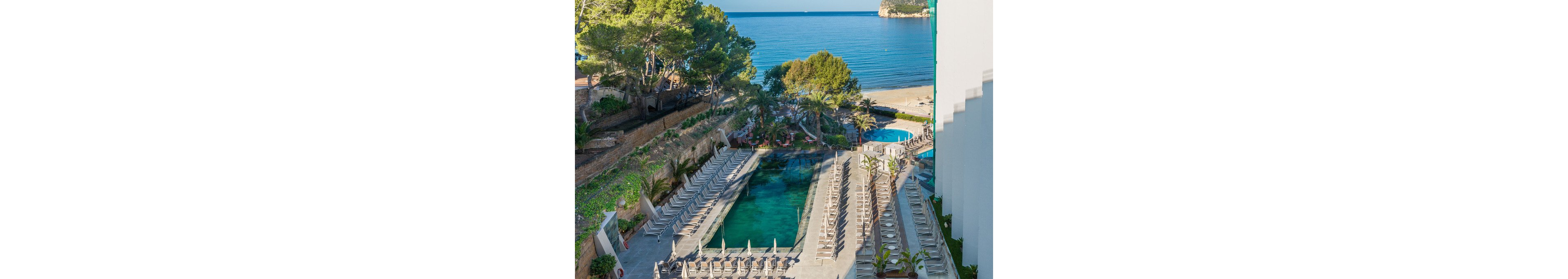 An elevated view shows a long rectangular hotel swimming pool lined with rows of empty sun loungers. The terrace is surrounded by trees, greenery, and a stone wall, with an additional circular pool visible near the sandy beach. Beyond the resort area, a calm blue sea stretches toward a rocky headland under clear skies. The scene suggests a tranquil vacation setting at a coastal hotel.