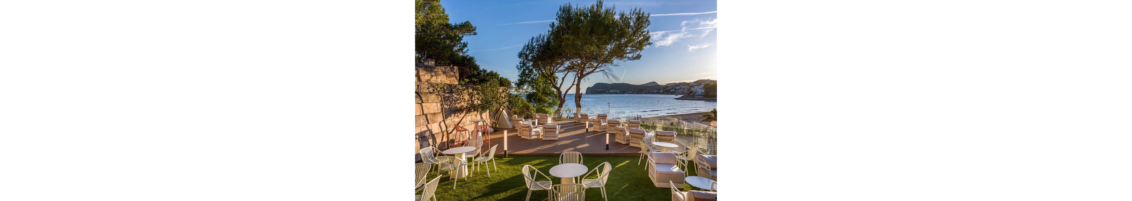 An outdoor terrace lounge with modern white chairs and sofas overlooks a calm seaside bay. The seating area is arranged on a wooden deck and adjacent green lawn, framed by rocky cliffs and lush trees. Gentle waves, a sandy beach, and distant hills are visible under a clear blue sky. Warm sunlight creates a relaxed, inviting atmosphere ideal for coastal dining or sunset drinks.