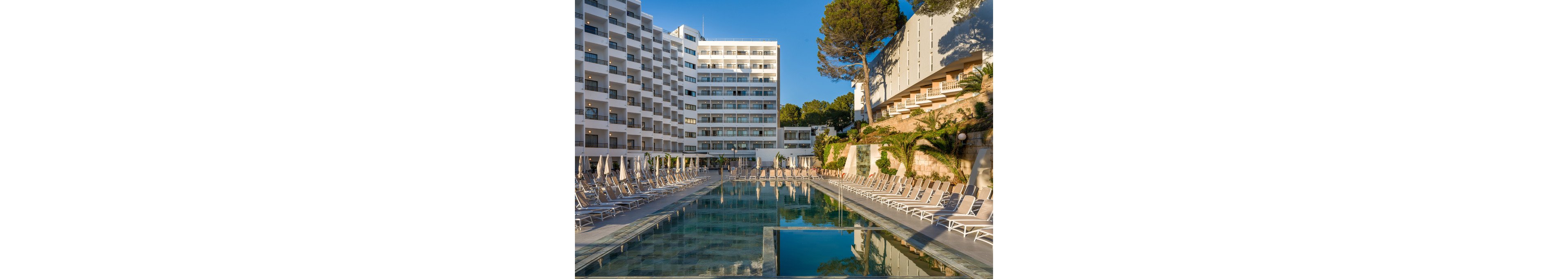 A large modern hotel complex overlooks a calm outdoor swimming pool lined with sun loungers and closed umbrellas. The scene is captured in warm daylight, highlighting the geometric facade of the multi-story building and the terraced structure on the right. Lush greenery and trees soften the architecture, creating a relaxed vacation atmosphere. The clear reflections in the pool emphasize the clean, contemporary resort setting.