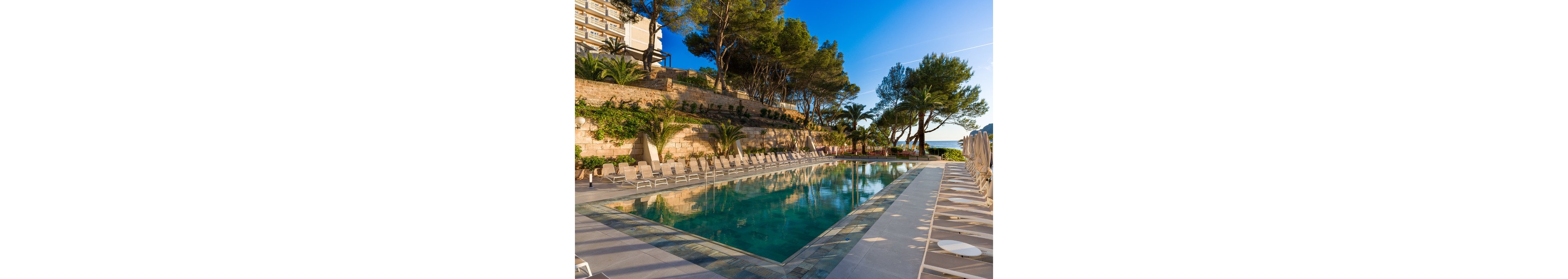 An outdoor swimming pool is surrounded by rows of beige lounge chairs at a hillside resort. Tall pine trees and stone retaining walls frame the pool area, creating a natural, Mediterranean feel. The clear blue water reflects the sky and nearby greenery, while a hotel building rises above the terrace. In the distance, the sea and coastline are visible under bright, warm sunlight.