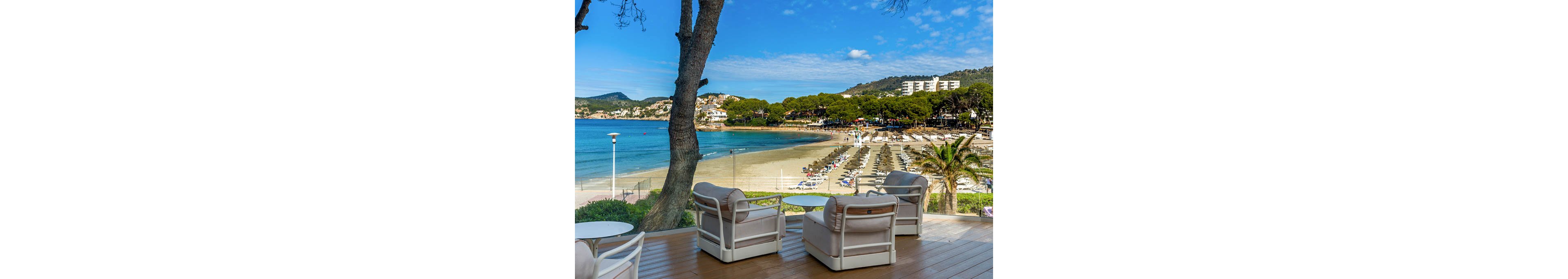 A seaside resort terrace with cushioned lounge chairs overlooks a curved sandy beach and calm turquoise water. Rows of sunbeds and umbrellas line the shoreline, suggesting a popular vacation destination. Pine trees, palm trees, and low-rise buildings frame the bay under a bright blue sky with scattered clouds. The scene conveys a relaxed, luxurious holiday atmosphere with a focus on outdoor leisure and coastal views.