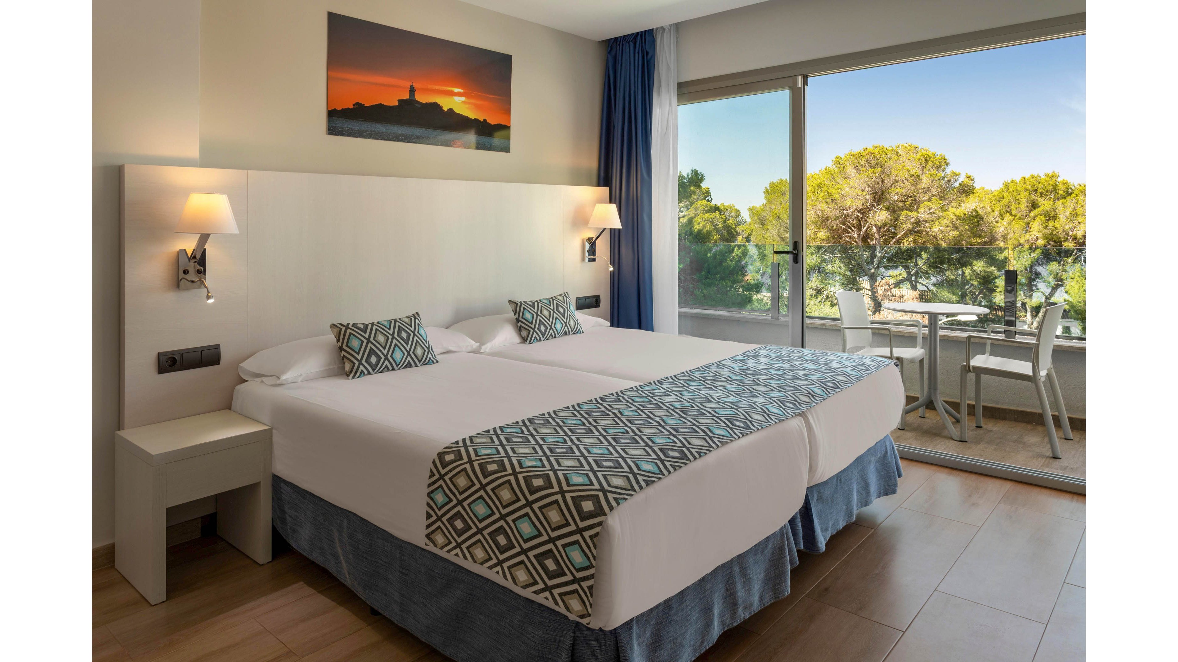 Contemporary hotel room featuring two neatly made twin beds with geometric patterned runners and cushions. The space is softly lit by wall-mounted lamps above minimalist bedside tables. A large glass sliding door opens to a balcony with outdoor seating and a scenic view of lush green trees. A framed sunset landscape photograph hangs above the headboard, adding warm color to the neutral interior.