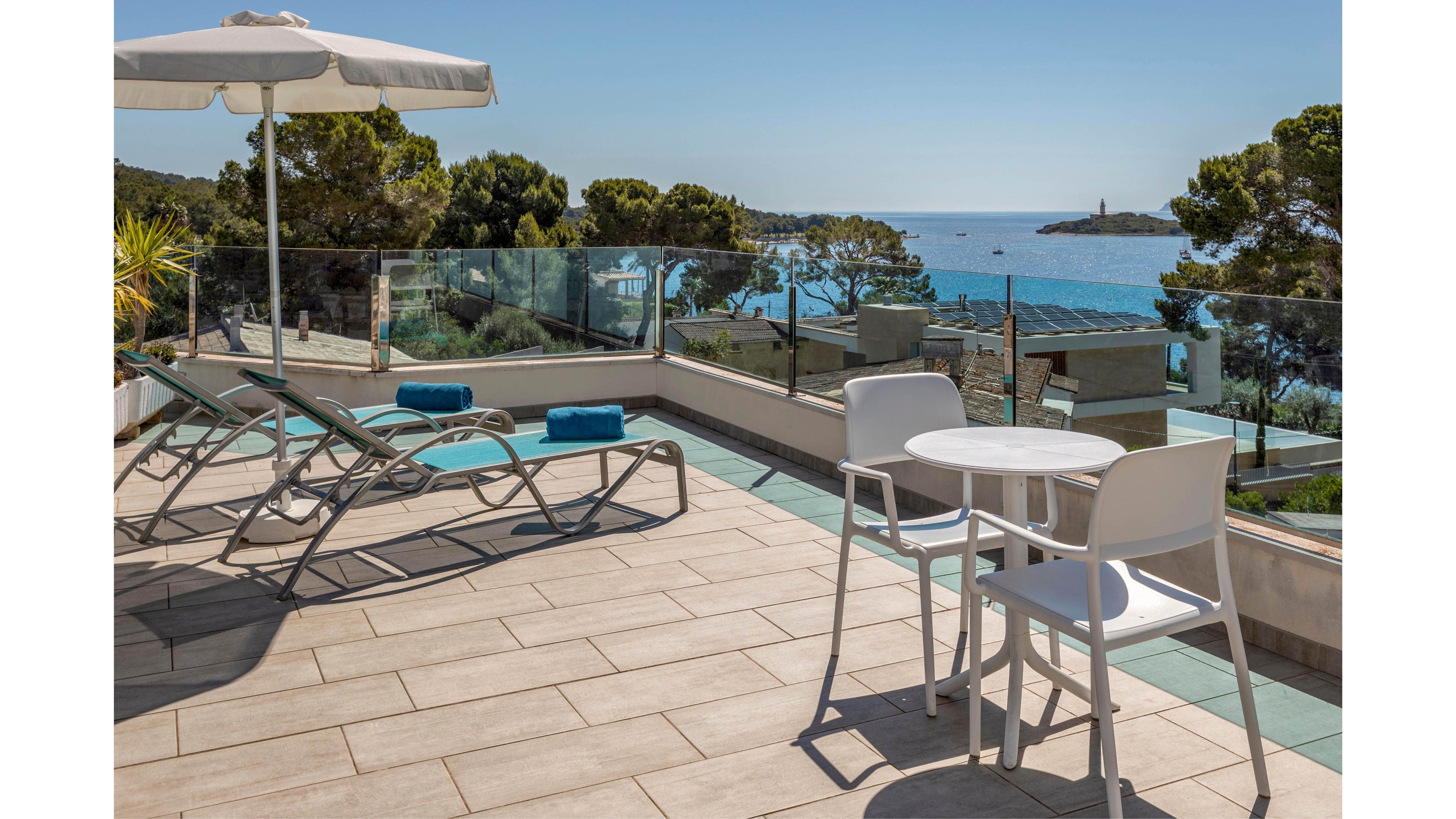 A modern terrace features white chairs and a small round table beside metal-framed sun loungers with blue cushions. A large white parasol provides shade on the tiled floor, while glass railings frame the view. Beyond the terrace, trees and coastal buildings lead down to a calm blue sea and distant rocky shoreline under a clear sky. The scene suggests a relaxing vacation setting with contemporary outdoor furniture and Mediterranean-style scenery.