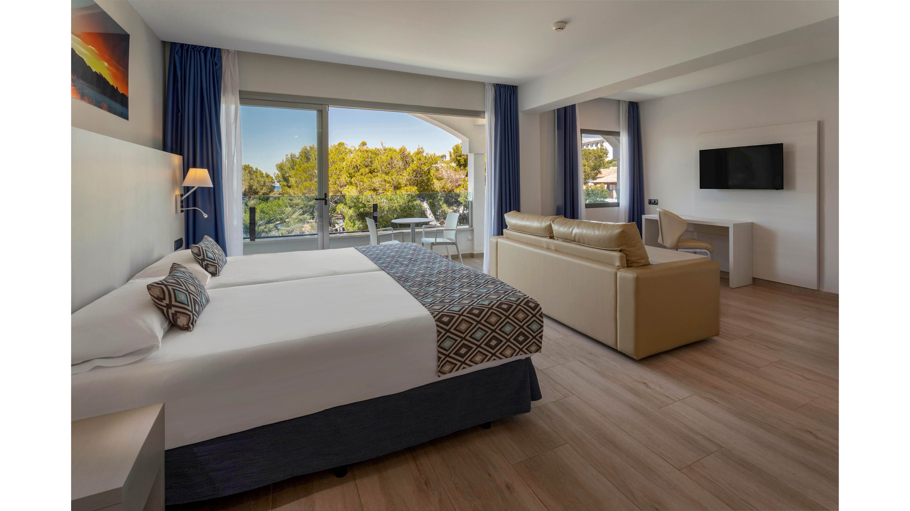 Spacious contemporary hotel room featuring two single beds with patterned cushions and a light wood floor. A sitting area with a beige sofa faces a wall-mounted flat-screen television. Large floor-to-ceiling windows open to a balcony overlooking green trees and nearby buildings. Blue curtains, neutral tones, and soft lighting create a calm, inviting atmosphere.