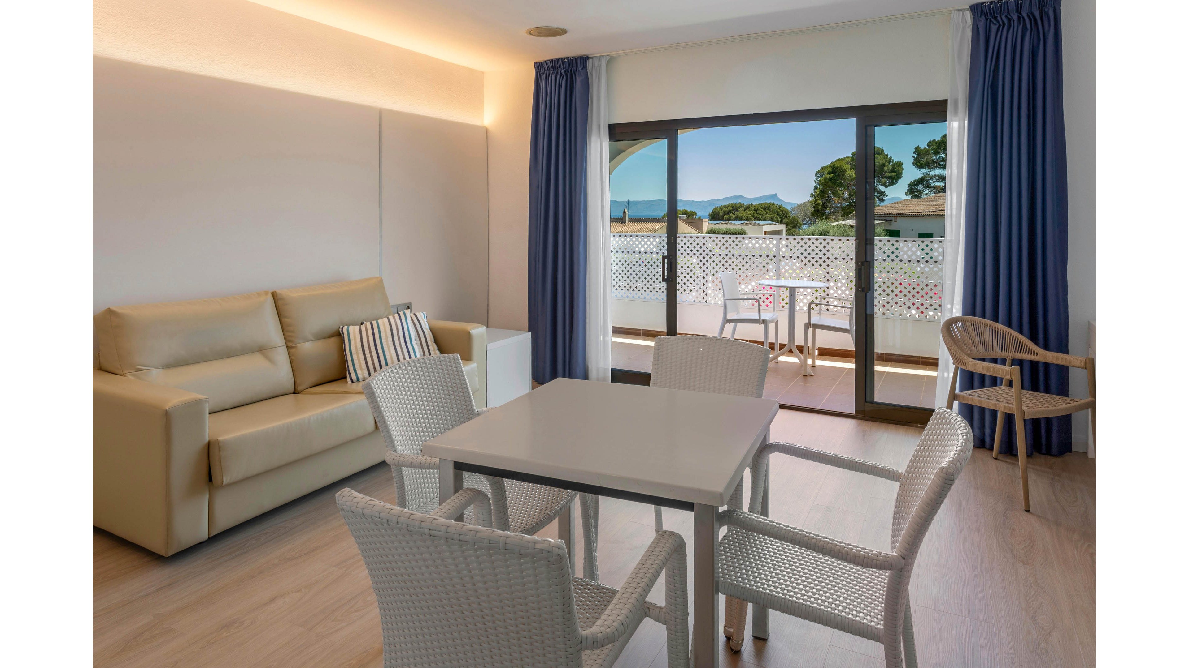 A contemporary living room features a beige sofa and a small dining table with four white woven chairs. Floor-to-ceiling glass doors open onto a balcony with outdoor seating and a scenic view of trees and distant hills. Blue curtains frame the doors, contrasting with the light wood flooring and neutral walls. Soft ambient lighting along the ceiling adds a warm, inviting atmosphere to the space.