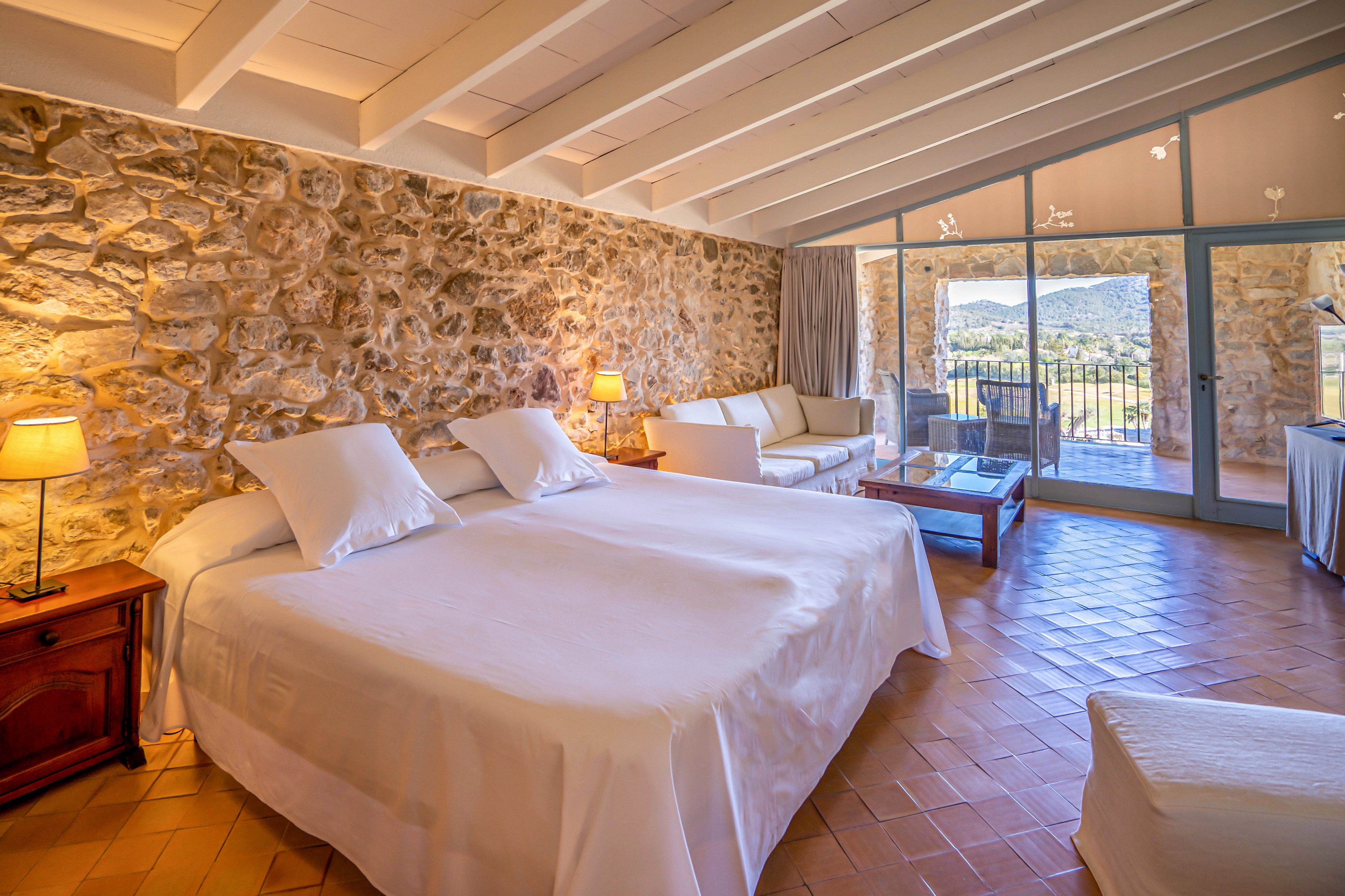 Bright, rustic-style hotel bedroom features a large double bed with white linens against a textured stone wall. The room includes bedside tables with warm lamps, a sofa seating area, and a coffee table. Floor-to-ceiling glass doors open to a terrace with outdoor furniture and a scenic mountain landscape. Exposed ceiling beams and terracotta floor tiles enhance the cozy, Mediterranean atmosphere.