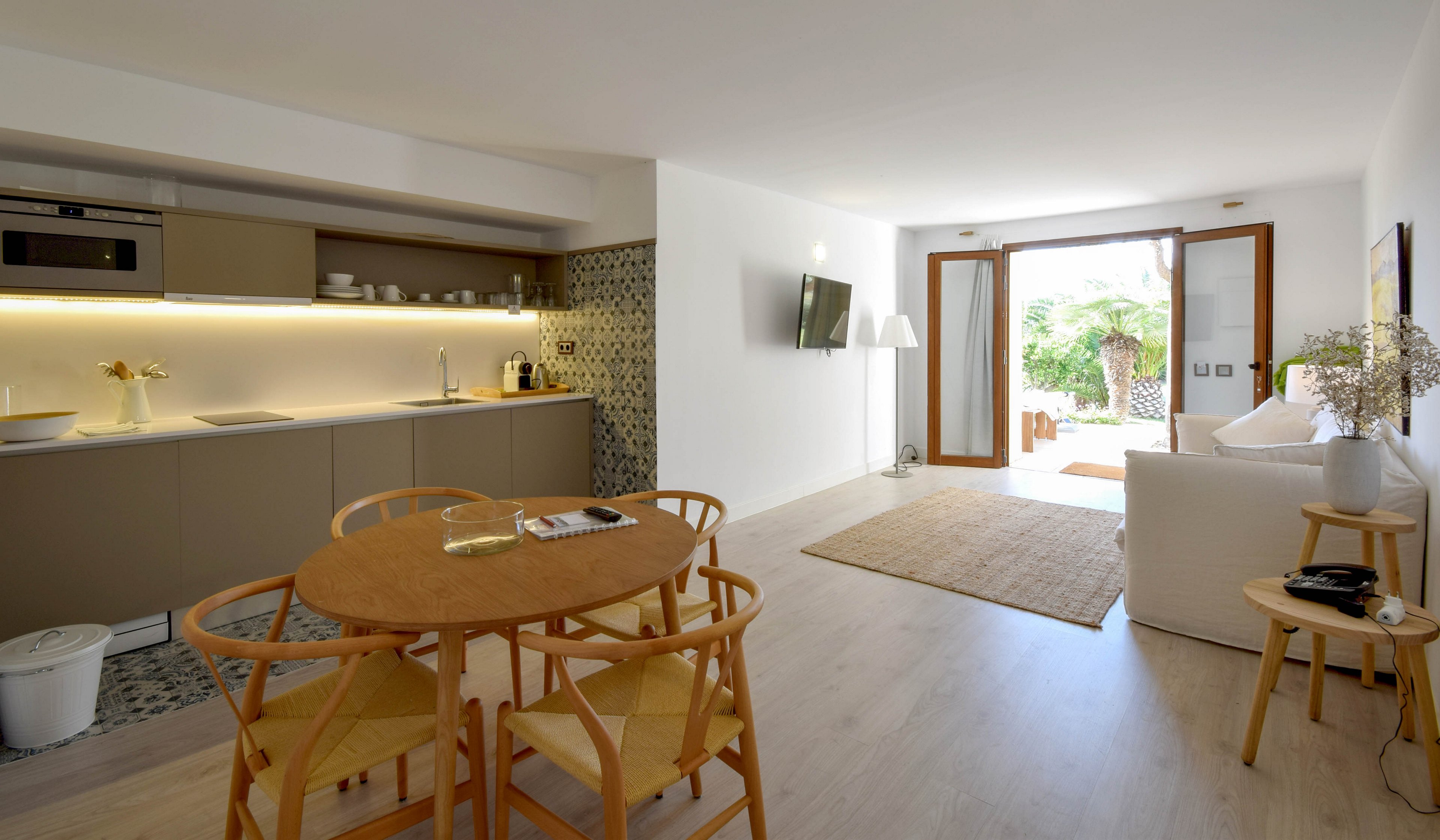 A contemporary open-plan studio apartment features a minimalist kitchenette and a small dining area with wooden chairs. The living space includes a white sofa, wall-mounted television, and neutral rug, all illuminated by natural light from large glass doors. Decorative elements such as plants, flowers, and patterned tiles add warmth and texture to the clean interior. Outside the open doors, greenery suggests a garden or patio area extending the living space.