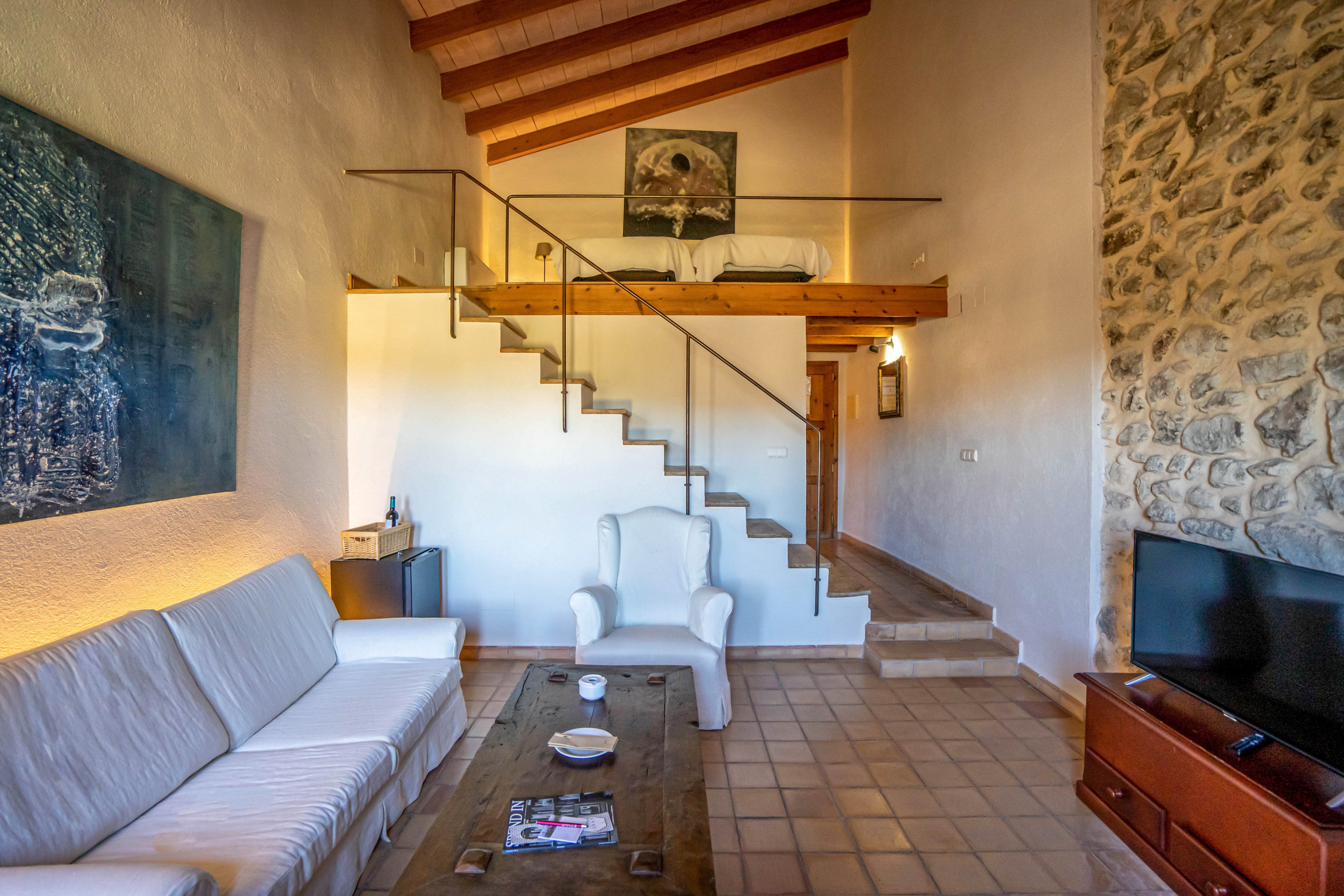 A bright living room features a white sofa and armchair around a wooden coffee table. A lofted sleeping area with a bed is accessed by a minimalist staircase with metal railing. Warm wooden ceiling beams and a stone accent wall create a rustic atmosphere. Soft lighting and wall art add a welcoming, homely feel to the space.