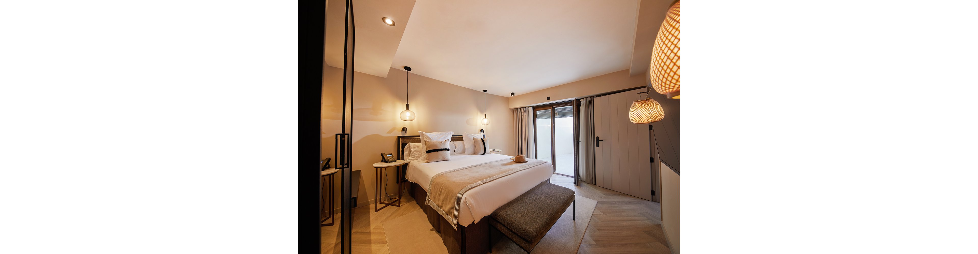 Contemporary hotel-style bedroom featuring a large double bed with neutral linens and accent pillows. Warm pendant lights hang above minimalist bedside tables, creating a soft and inviting atmosphere. A cushioned bench sits at the foot of the bed, and a wide glass door with curtains opens to an outdoor area. The room is decorated in beige and brown tones with clean lines and subtle modern details.